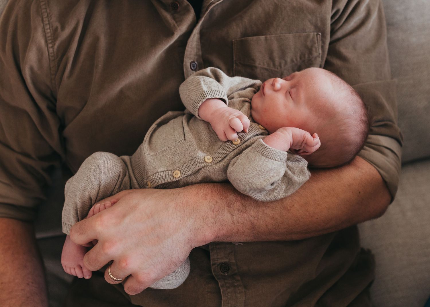 Baby sleeping peacefully in grey onesie while resting on parent's arms.
