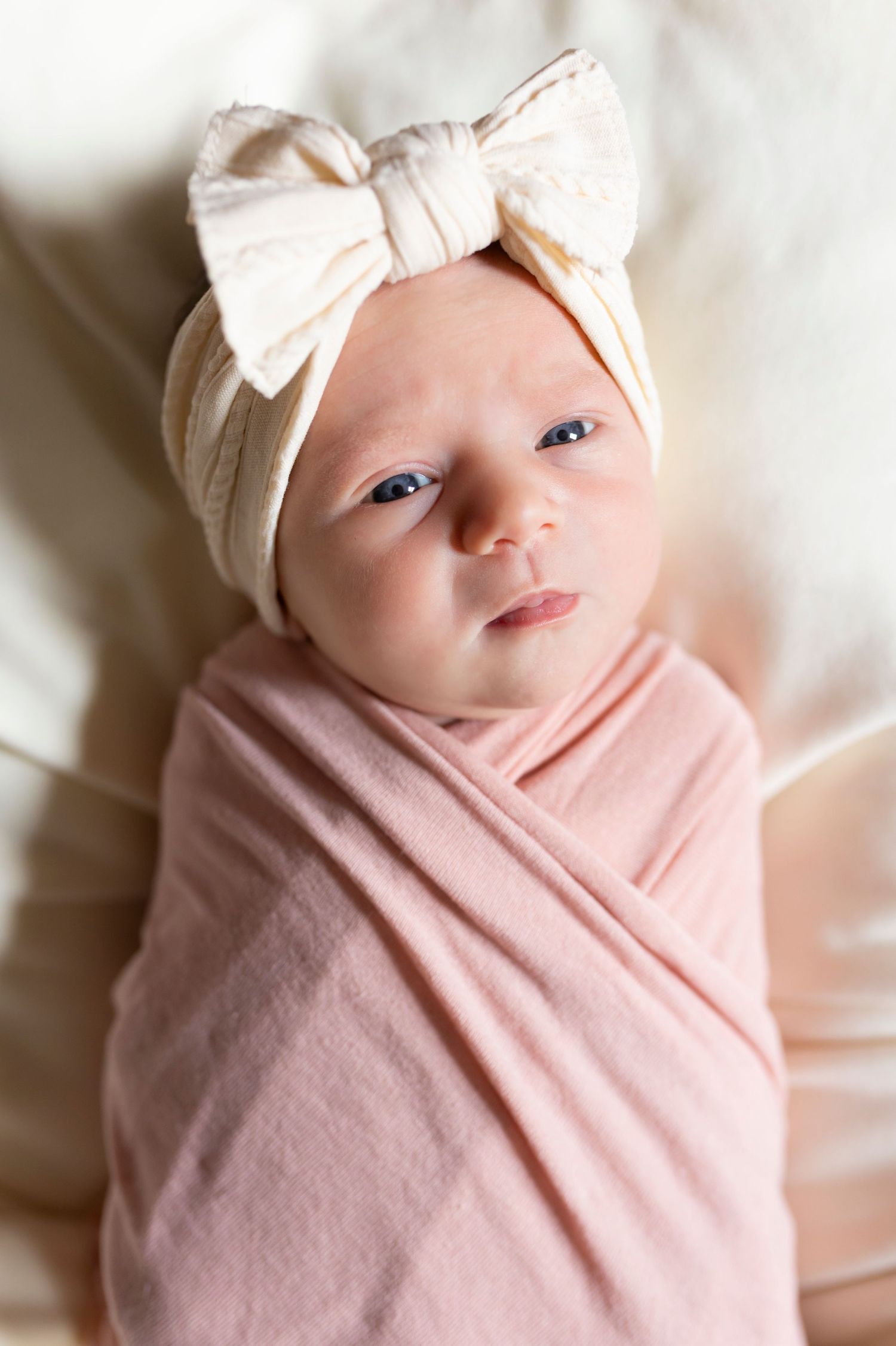 Newborn baby swaddled in pink wrap with white bow headband smiling at camera.