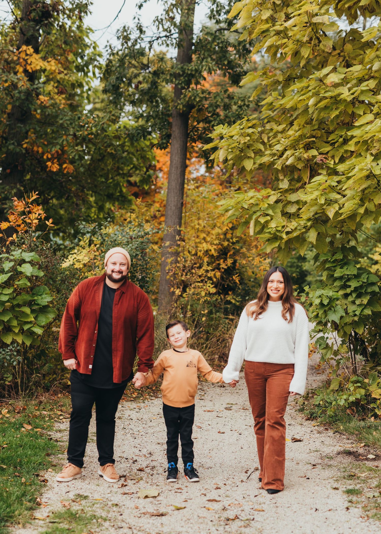 A family walks together on a leaf-covered path through autumn trees with golden foliage on a sunny day.