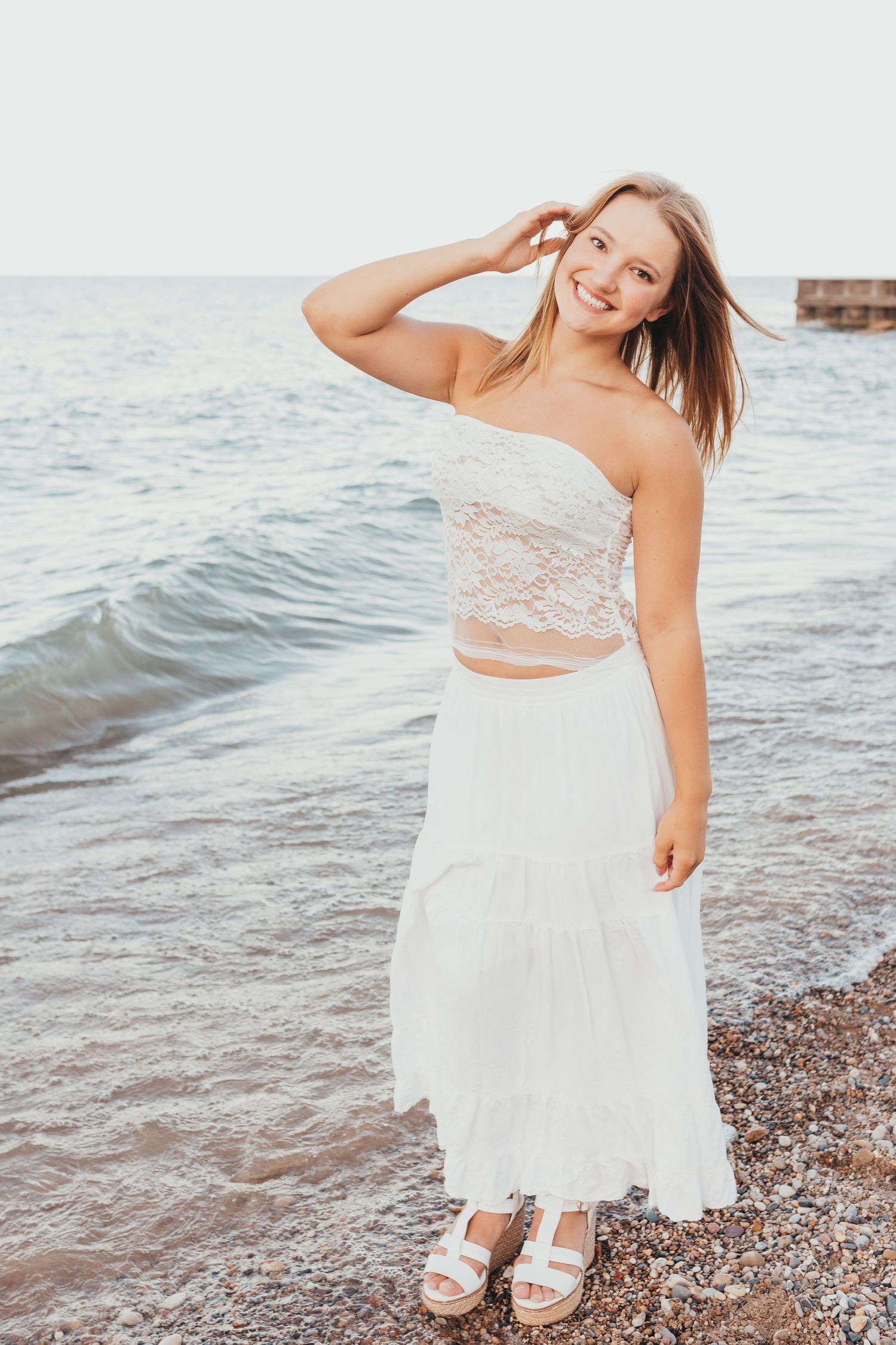 A white lace dress flows elegantly against a beach backdrop with gentle waves and rocky shore.