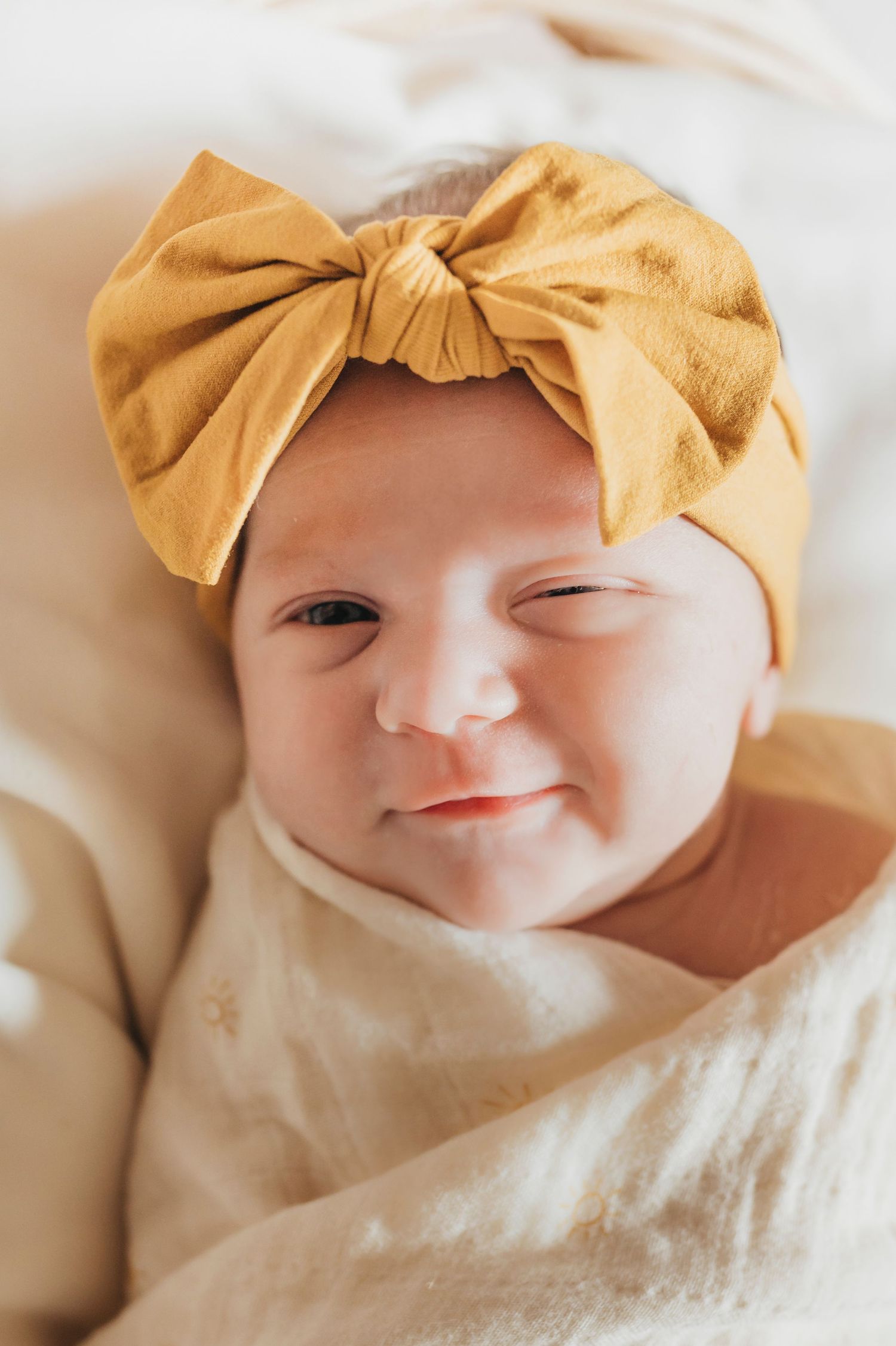 Baby wearing yellow headband smiles sweetly while wrapped in white blanket.