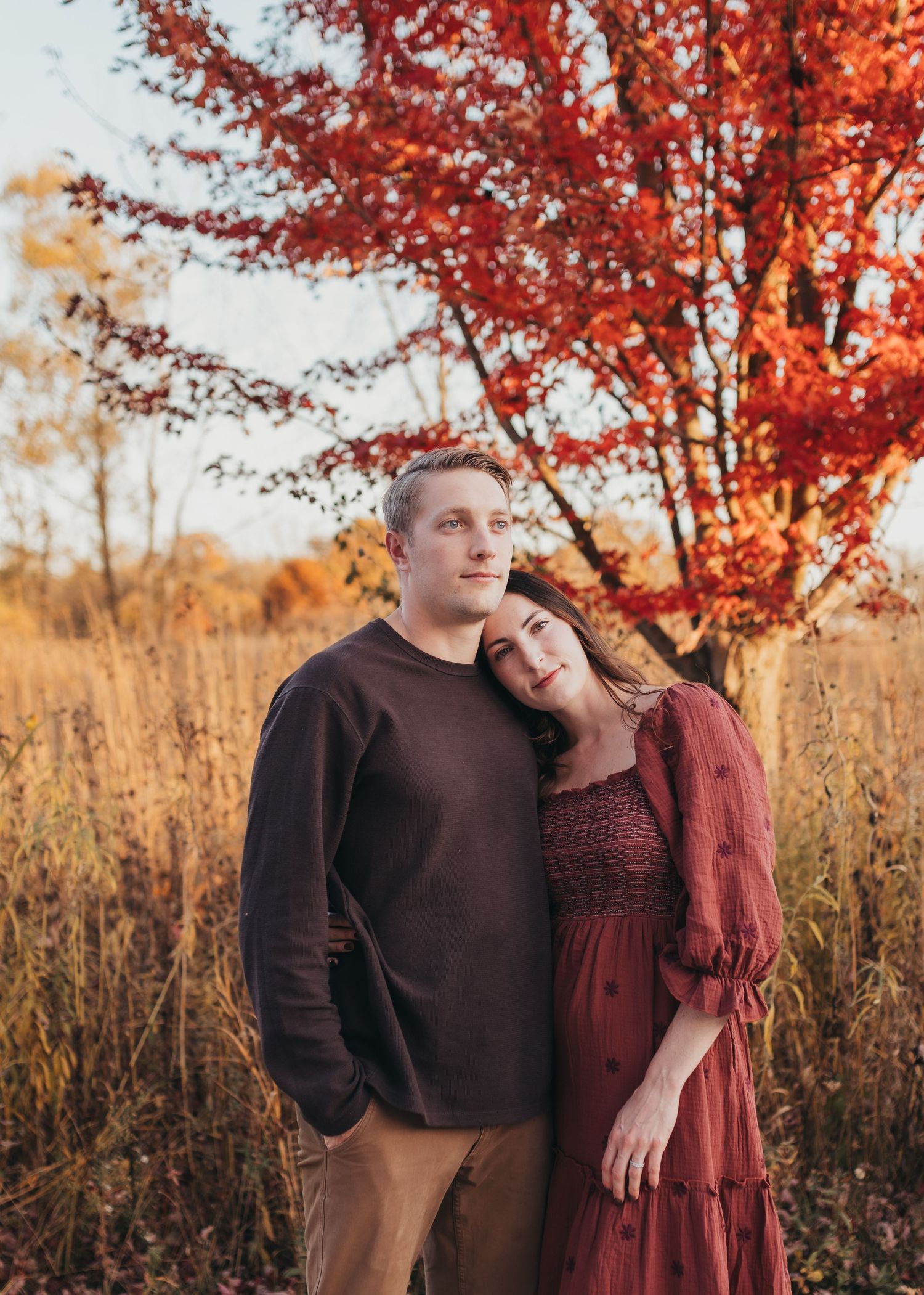 A couple embraces under vibrant red autumn trees in a golden field during sunset.