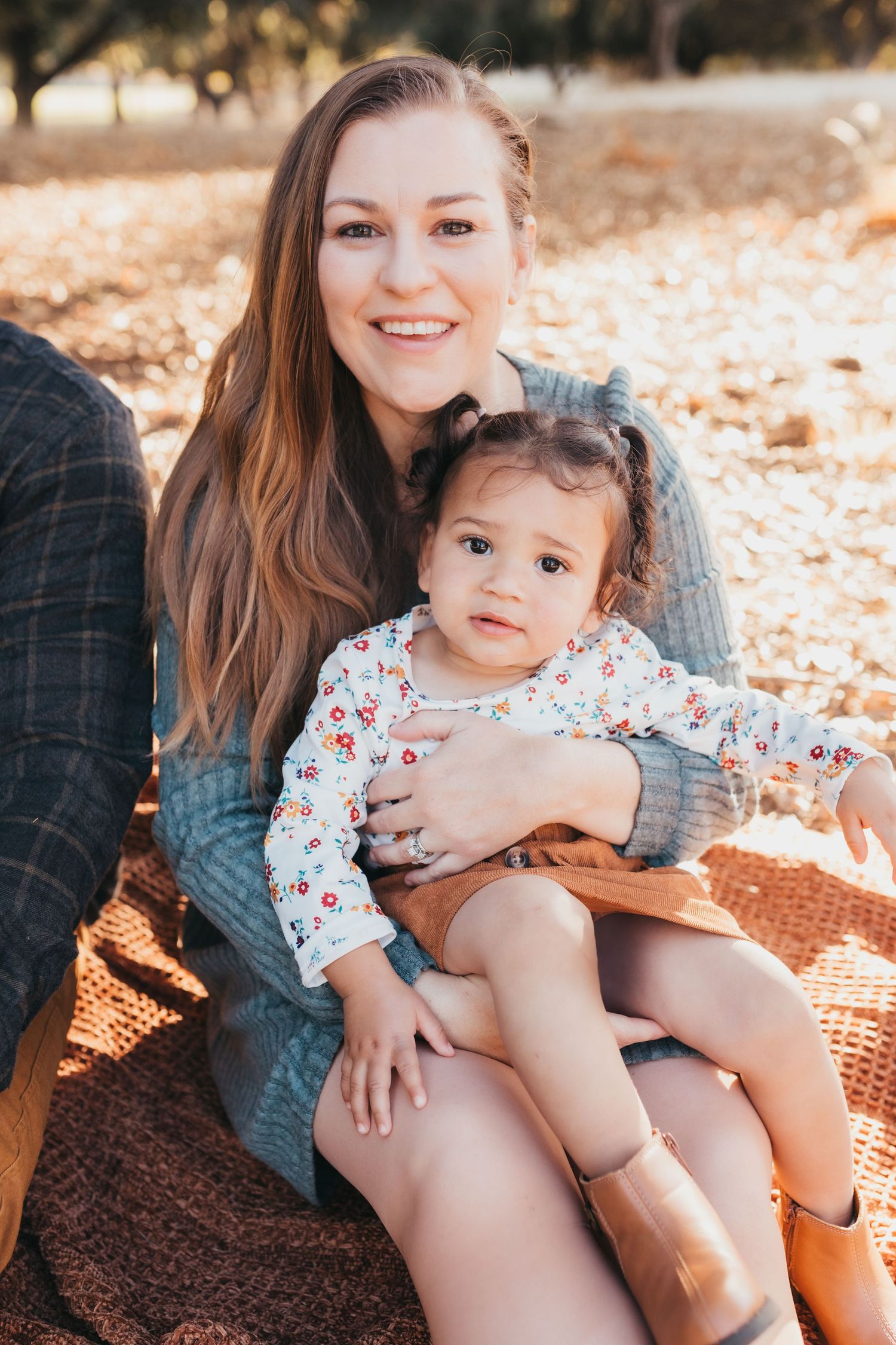 A parent and young child sit together on a blanket outdoors during autumn with fallen leaves in the background.