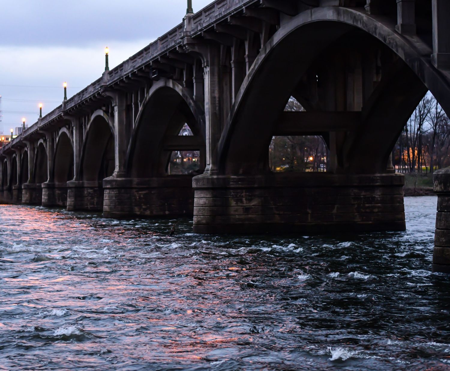Stone arch bridge spans across water at dusk with dramatic lighting and reflections.