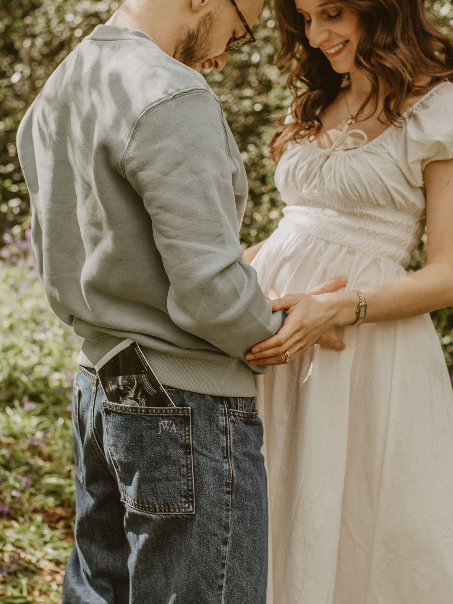 Pregnant woman in white dress with partner holding her baby bump outdoors, ultrasound photo in his pocket.