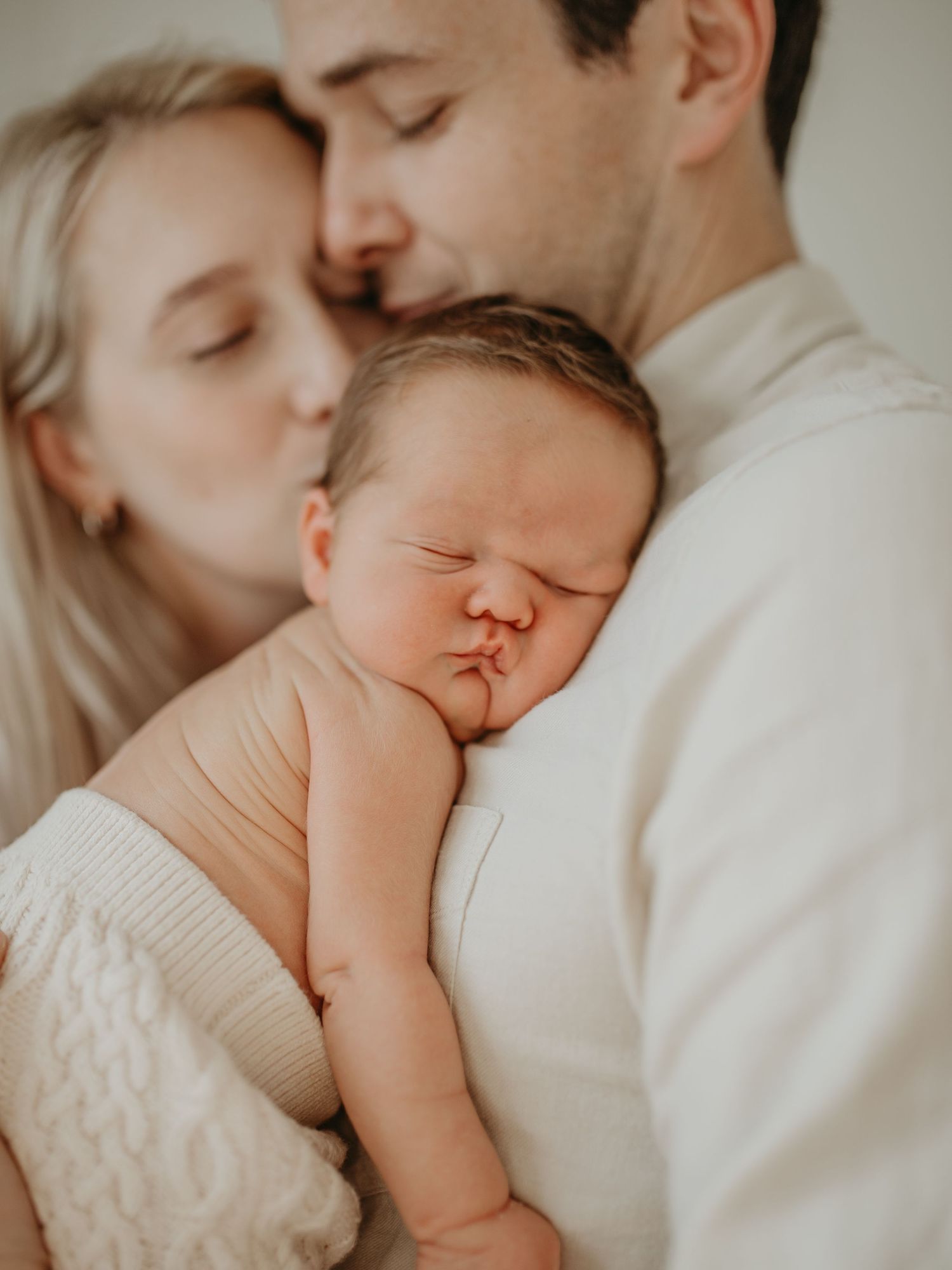 Tender family moment showing parent and sleeping baby cuddled close together on white bedding in soft natural light.