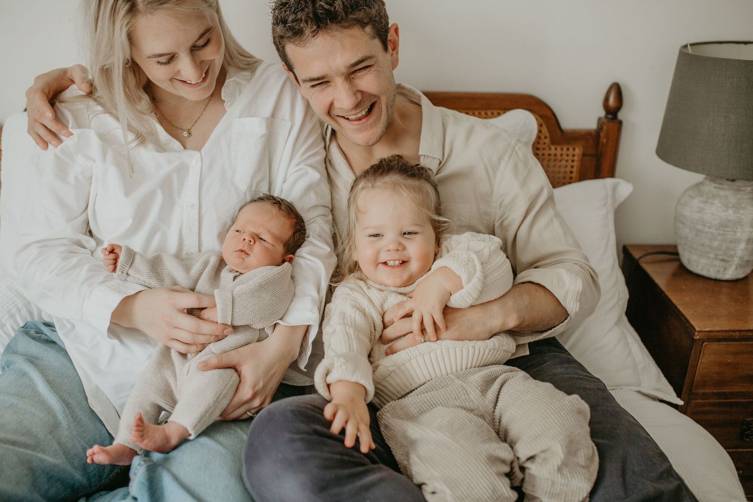 A family relaxes together on a bed, with a newborn baby and toddler in cozy knit outfits.