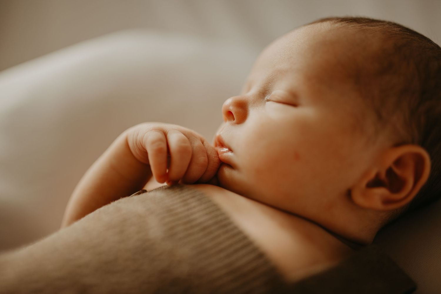 A peaceful sleeping newborn baby in warm light rests with tiny hand curled near their face.