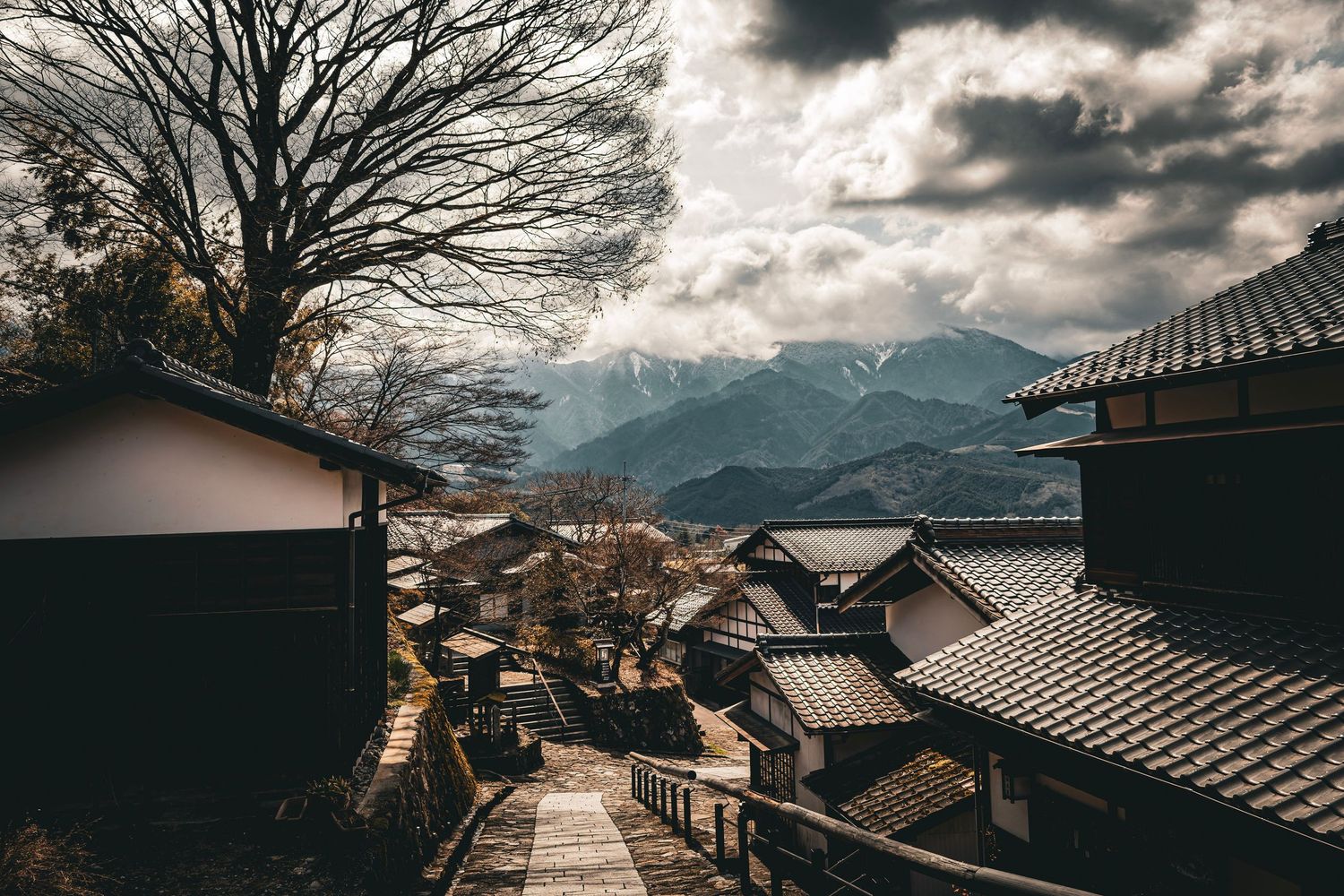 Traditional Japanese village street with historic wooden buildings, bare trees, and snow-capped mountains under dramatic clouds.