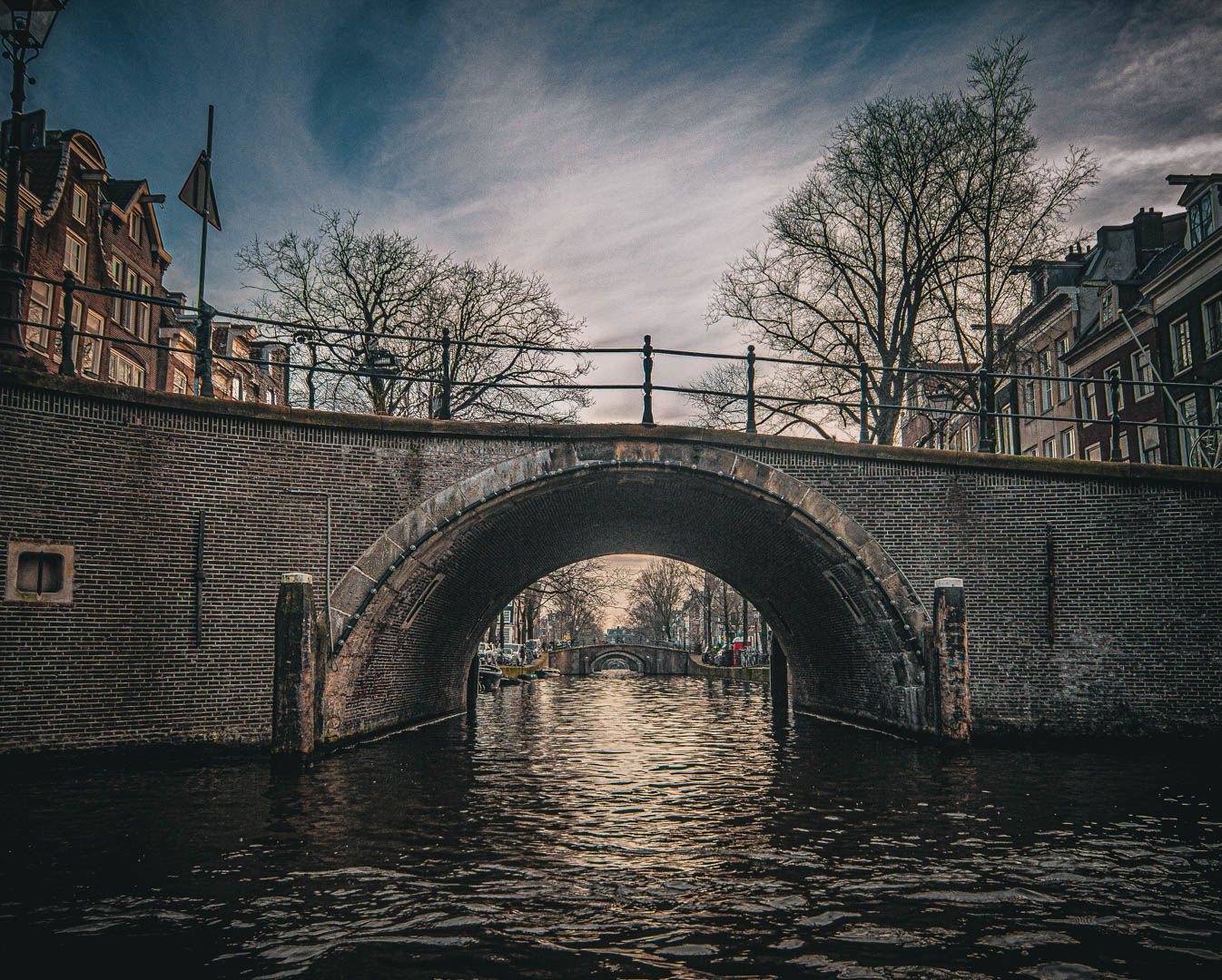 Historic brick arch bridge spanning an Amsterdam canal under dramatic cloudy skies in winter.