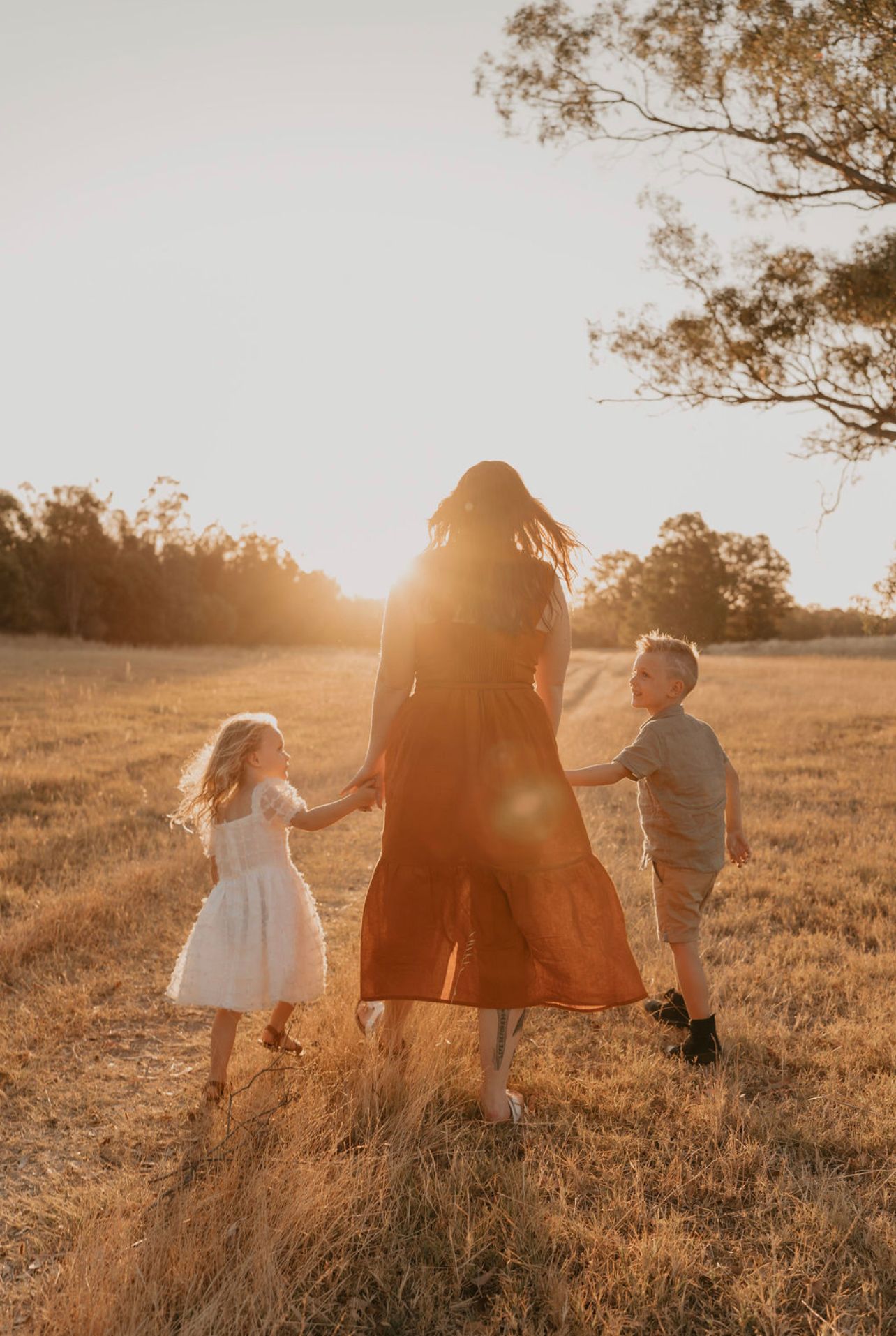 Mother and two young children walking hand in hand through golden sunlit field at sunset.