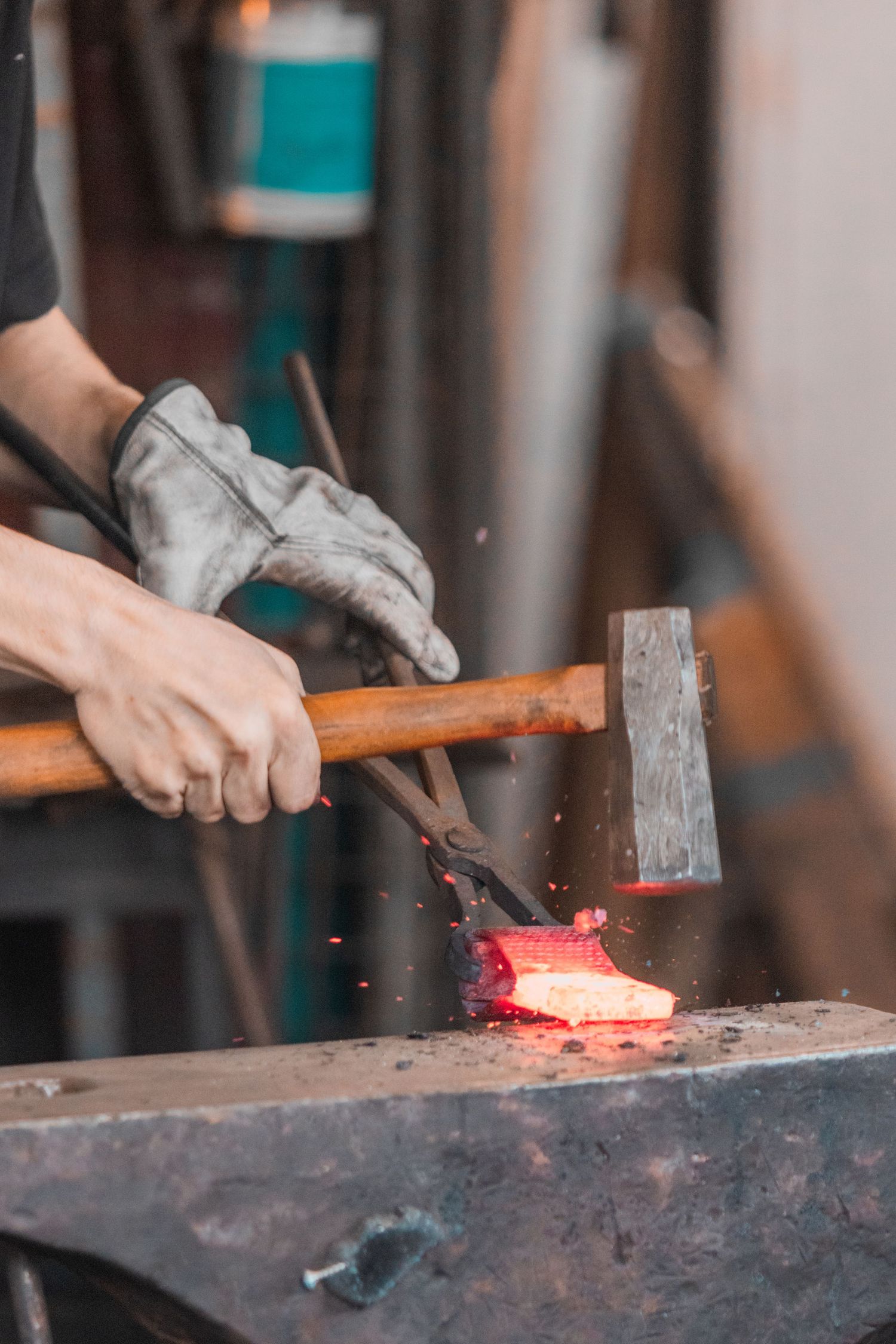 A blacksmith hammering glowing red-hot metal on an anvil with tongs, wearing protective gloves in a forge workshop.