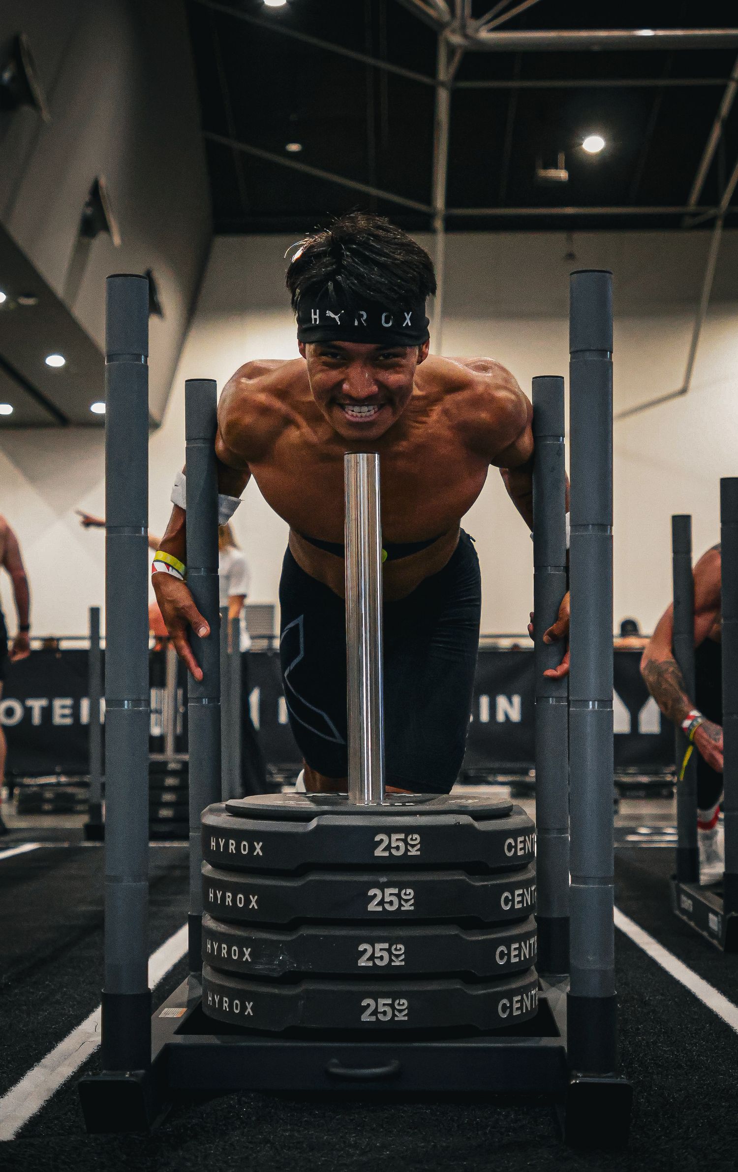 Muscular shirtless athlete pushes a weighted HYROX sled during an intense fitness competition in a gym.