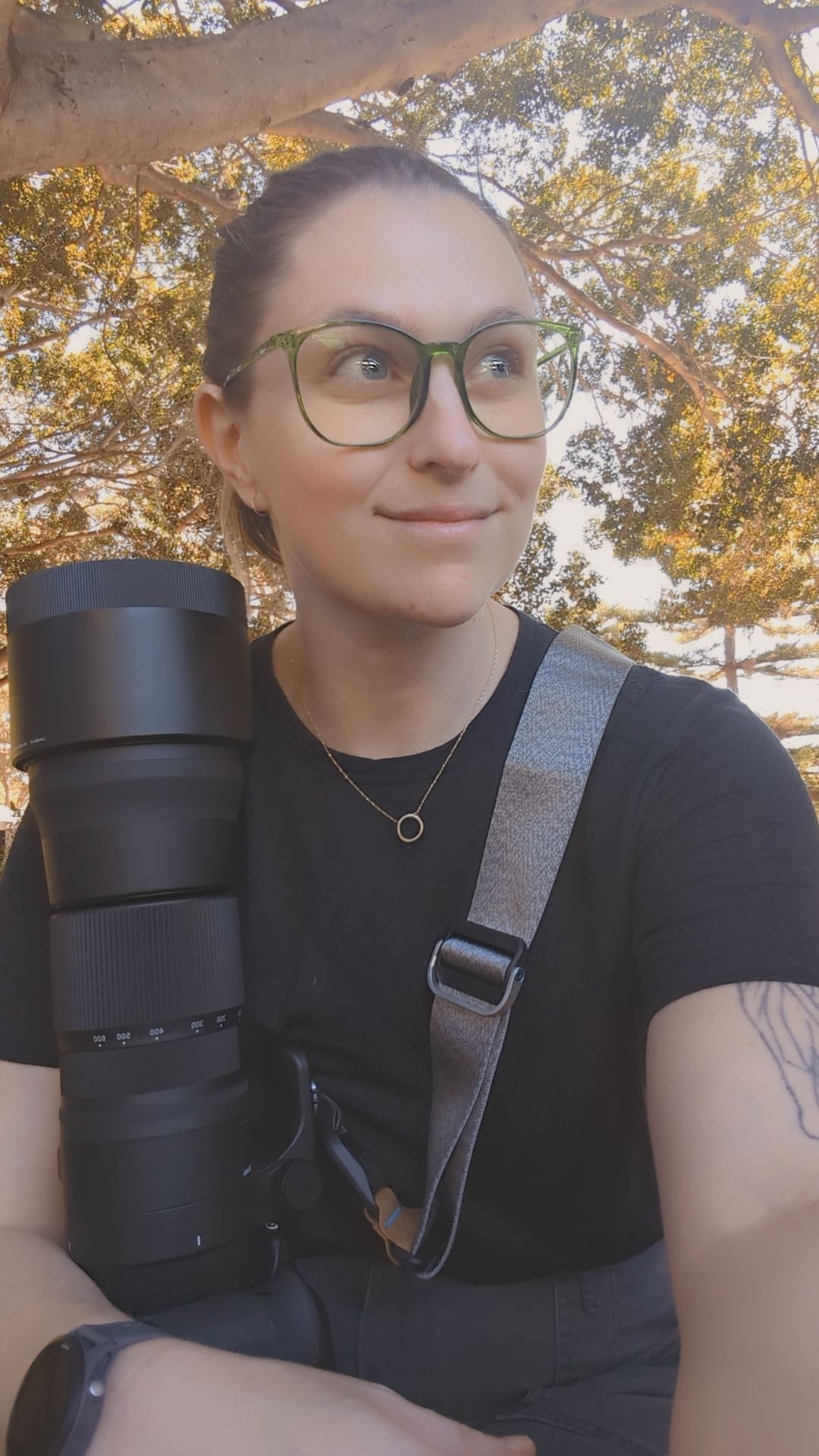 Young woman with green glasses holding a large telephoto camera lens outdoors under a tree.