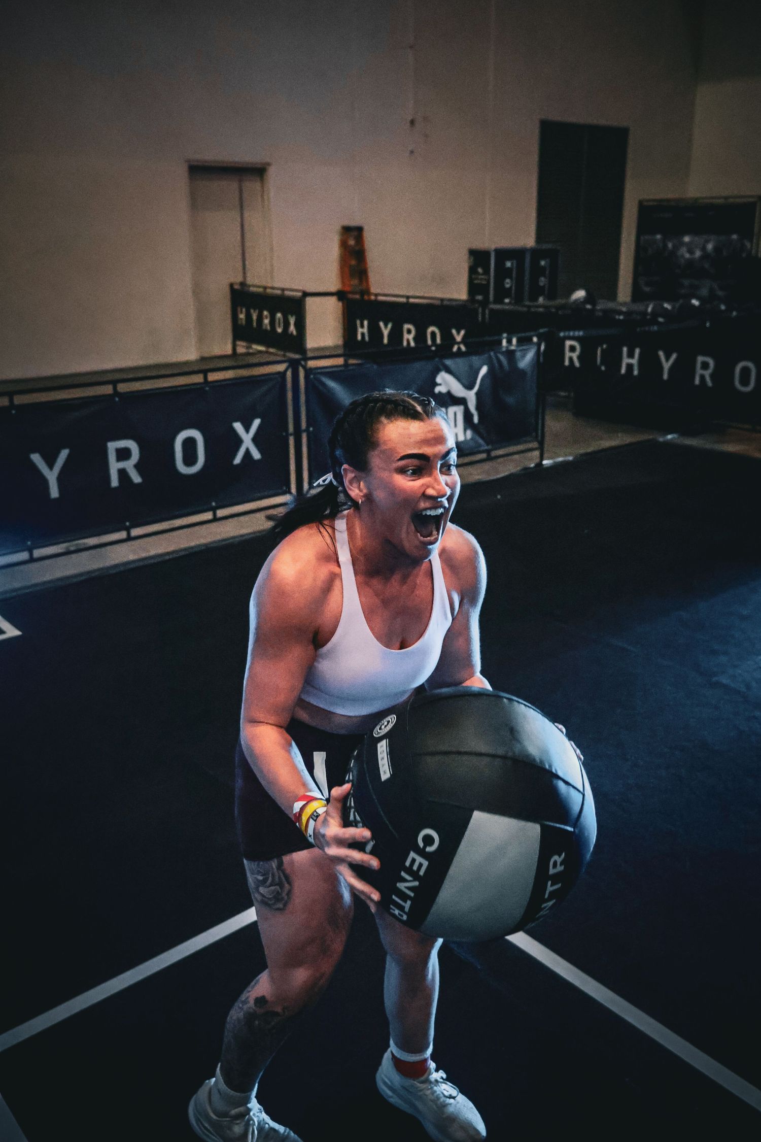 Athlete intensely holds a medicine ball during a HYROX fitness competition in a dark indoor gym.