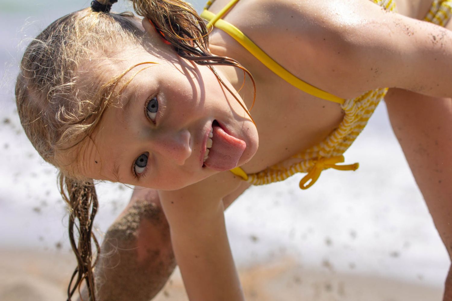 A young girl in a yellow bikini at the beach, sticking out her tongue and smiling playfully.