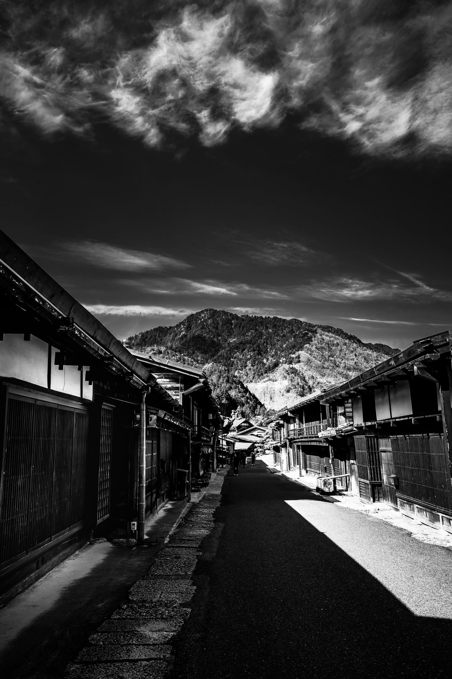 Black and white photo of a quiet traditional Japanese street flanked by old wooden buildings and a mountain backdrop.