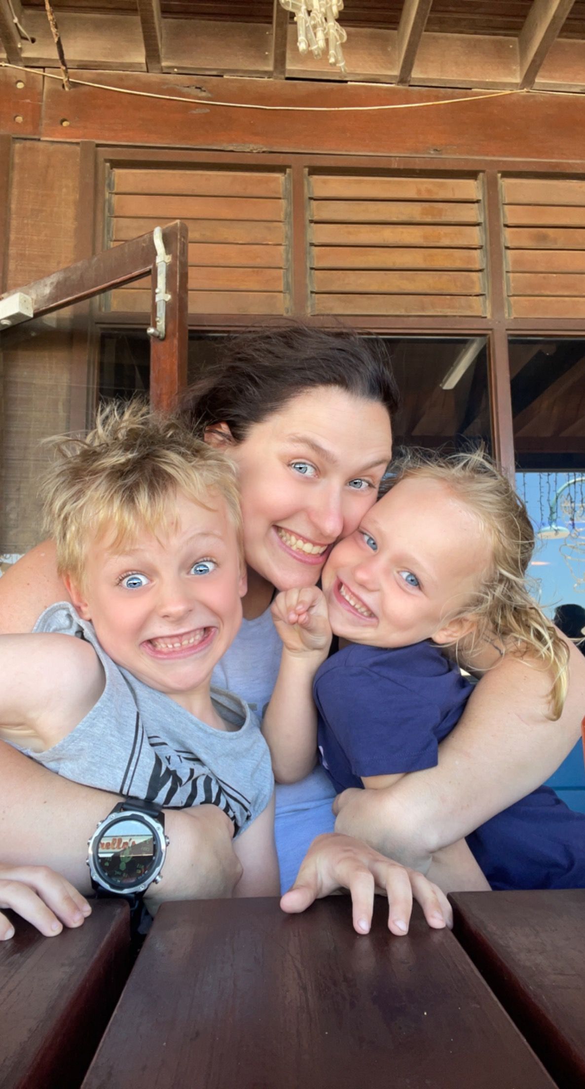 Happy woman and two blonde blue-eyed children smiling together at a wooden outdoor table.