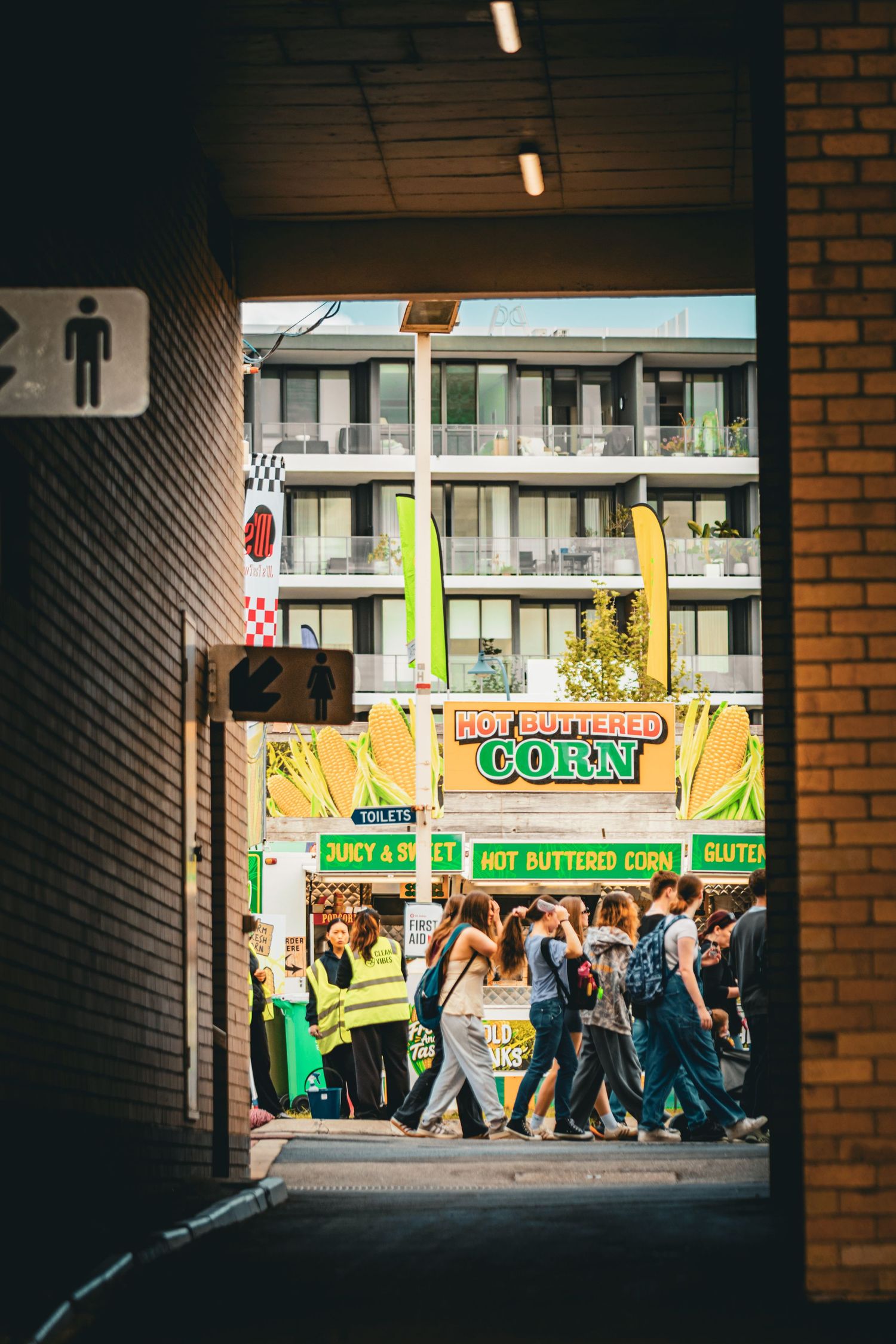 View through a tunnel of a busy street fair with a Hot Buttered Corn food stall and crowds of people outside.