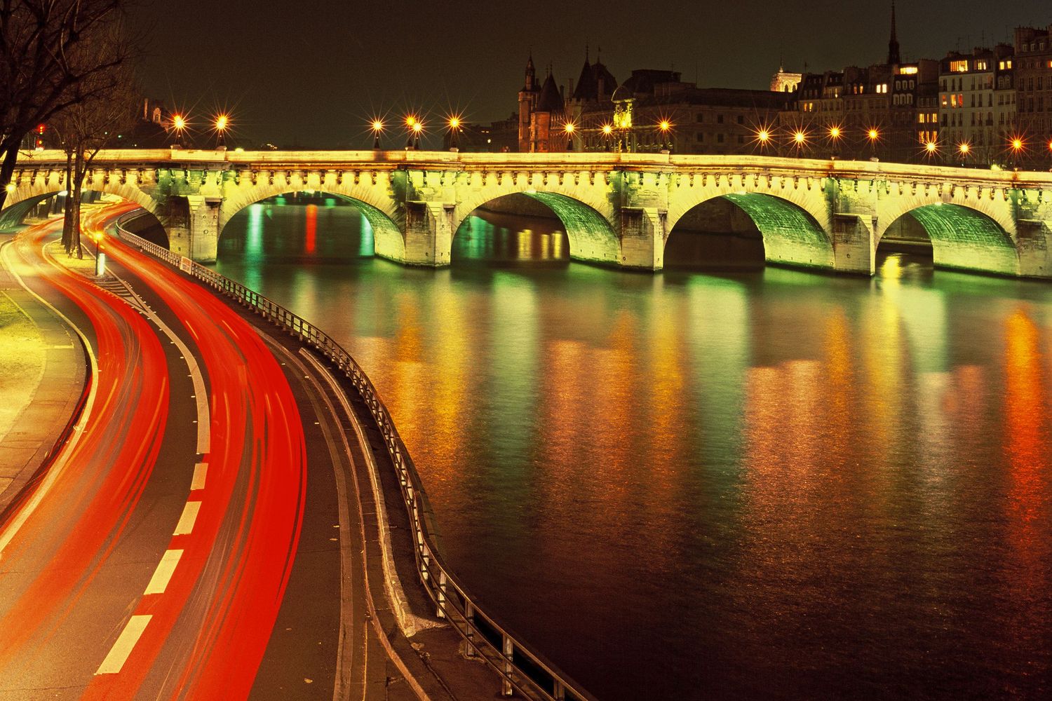 Long-exposure night view of illuminated Pont Neuf and red traffic trails beside the Seine in Paris.