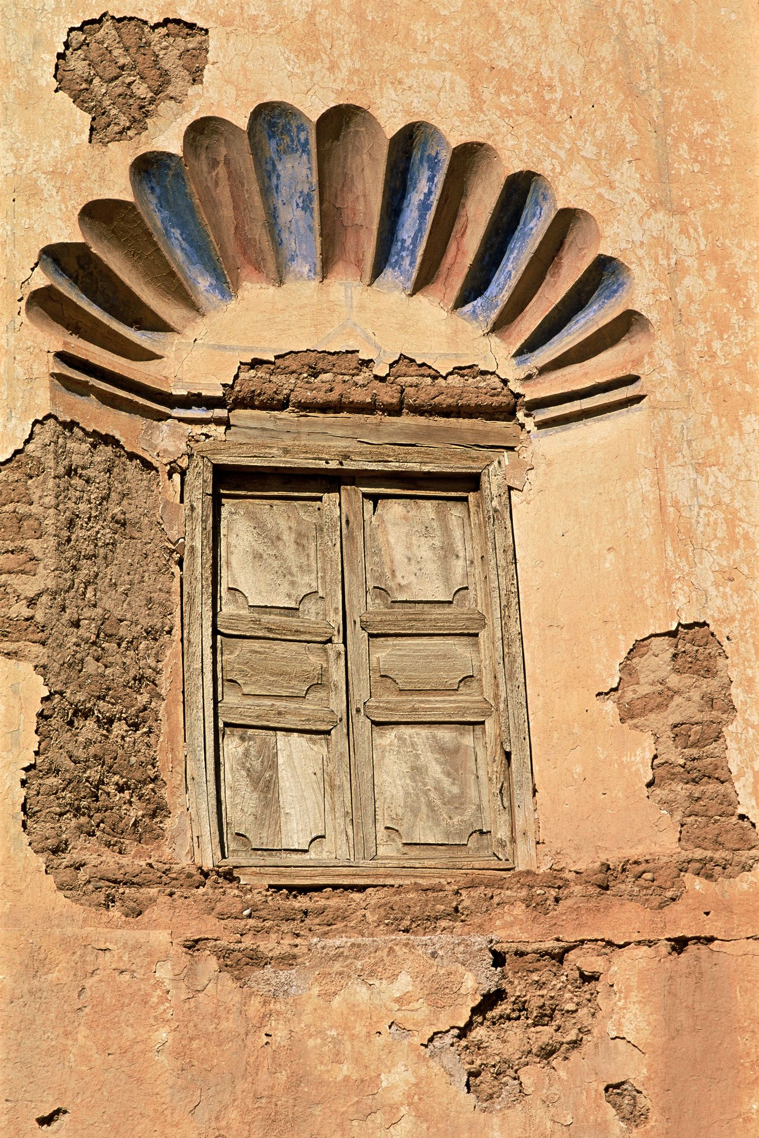 Weathered wooden window set in a crumbling ochre wall beneath a decorative arch in Morocco.