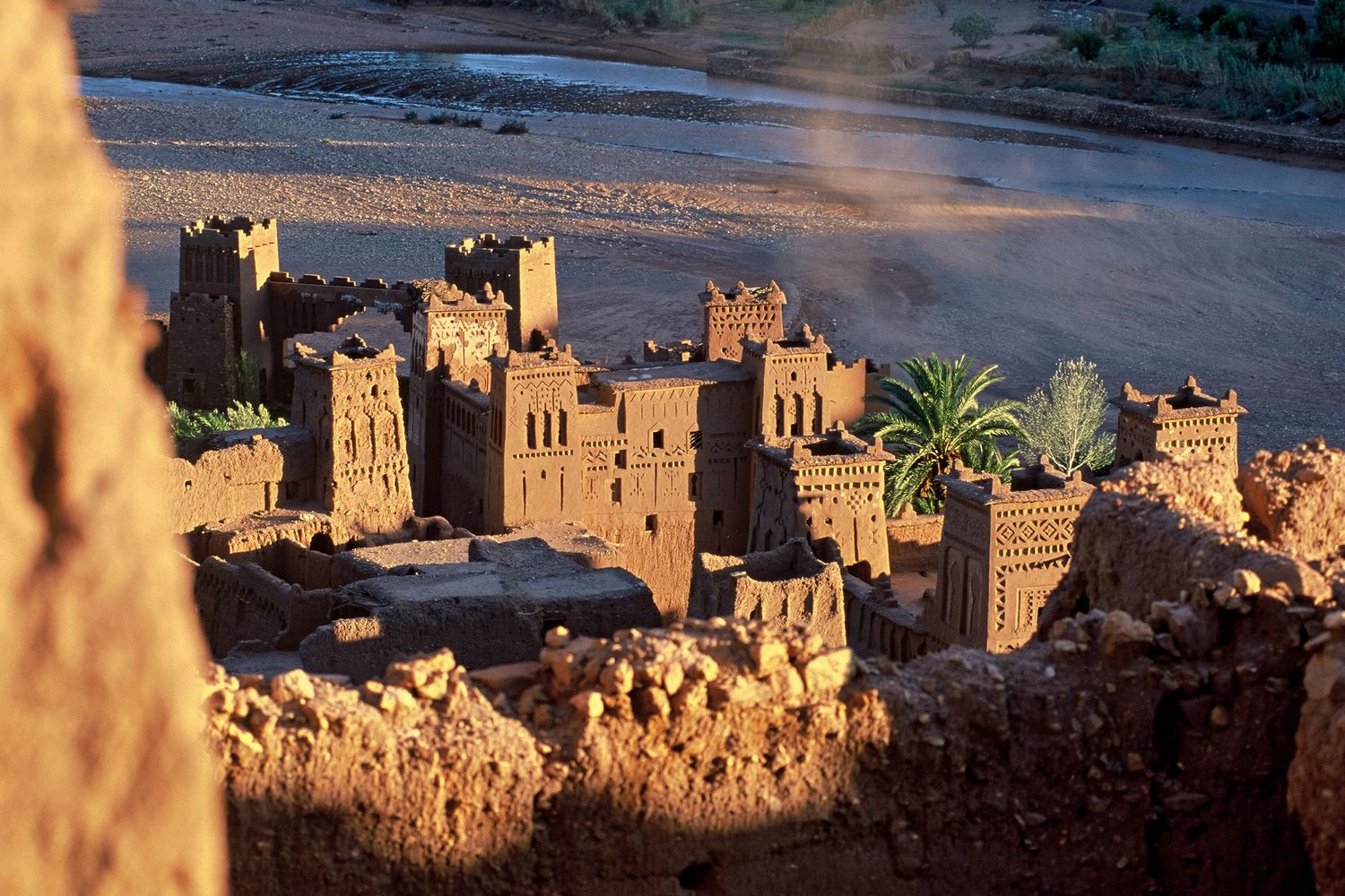 Sunlit earthen buildings of Aït Benhaddou rising above a river valley in Morocco.