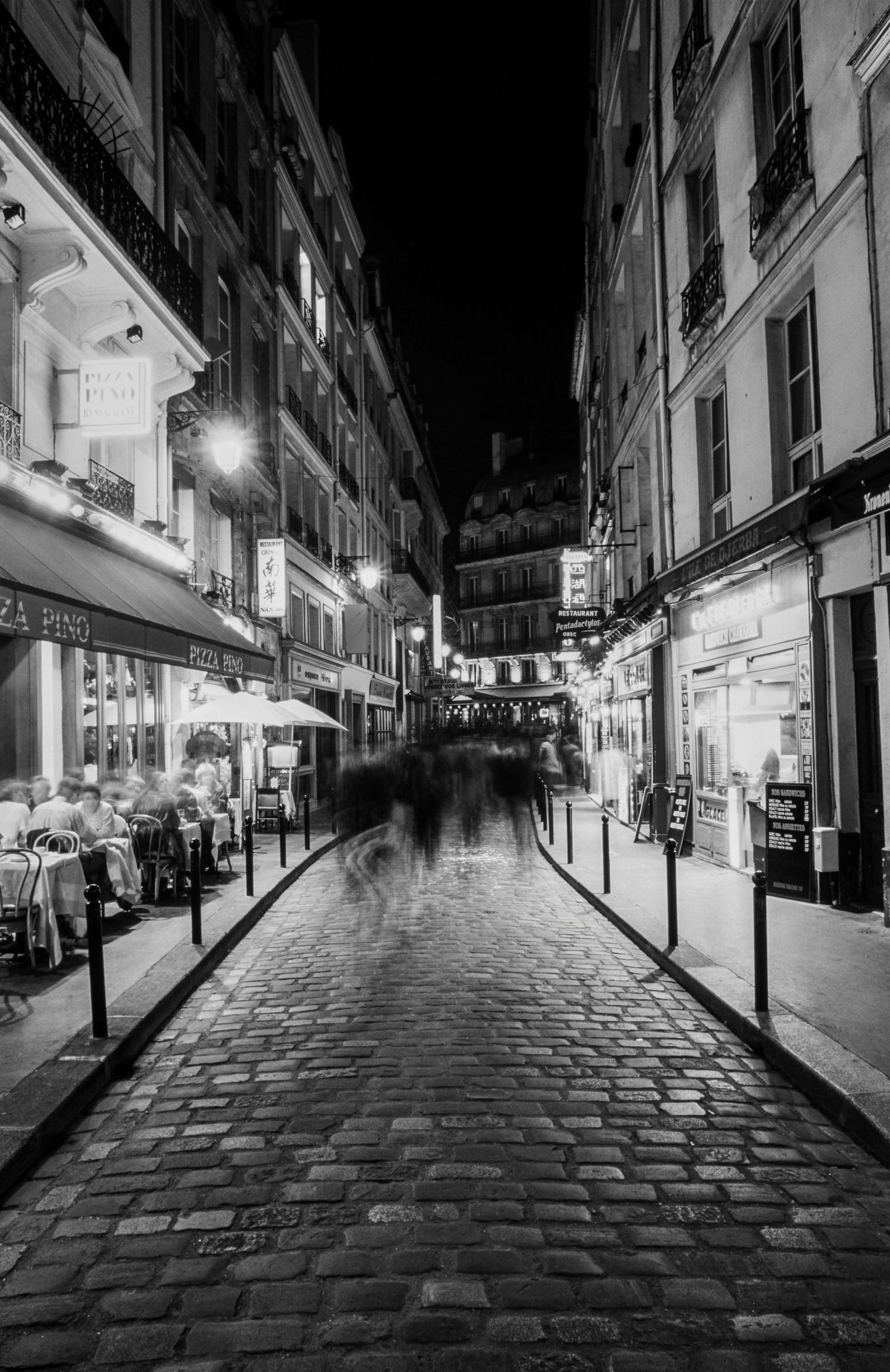 Black-and-white view of a cobblestone street lined with cafés and buildings at night in Paris, France.