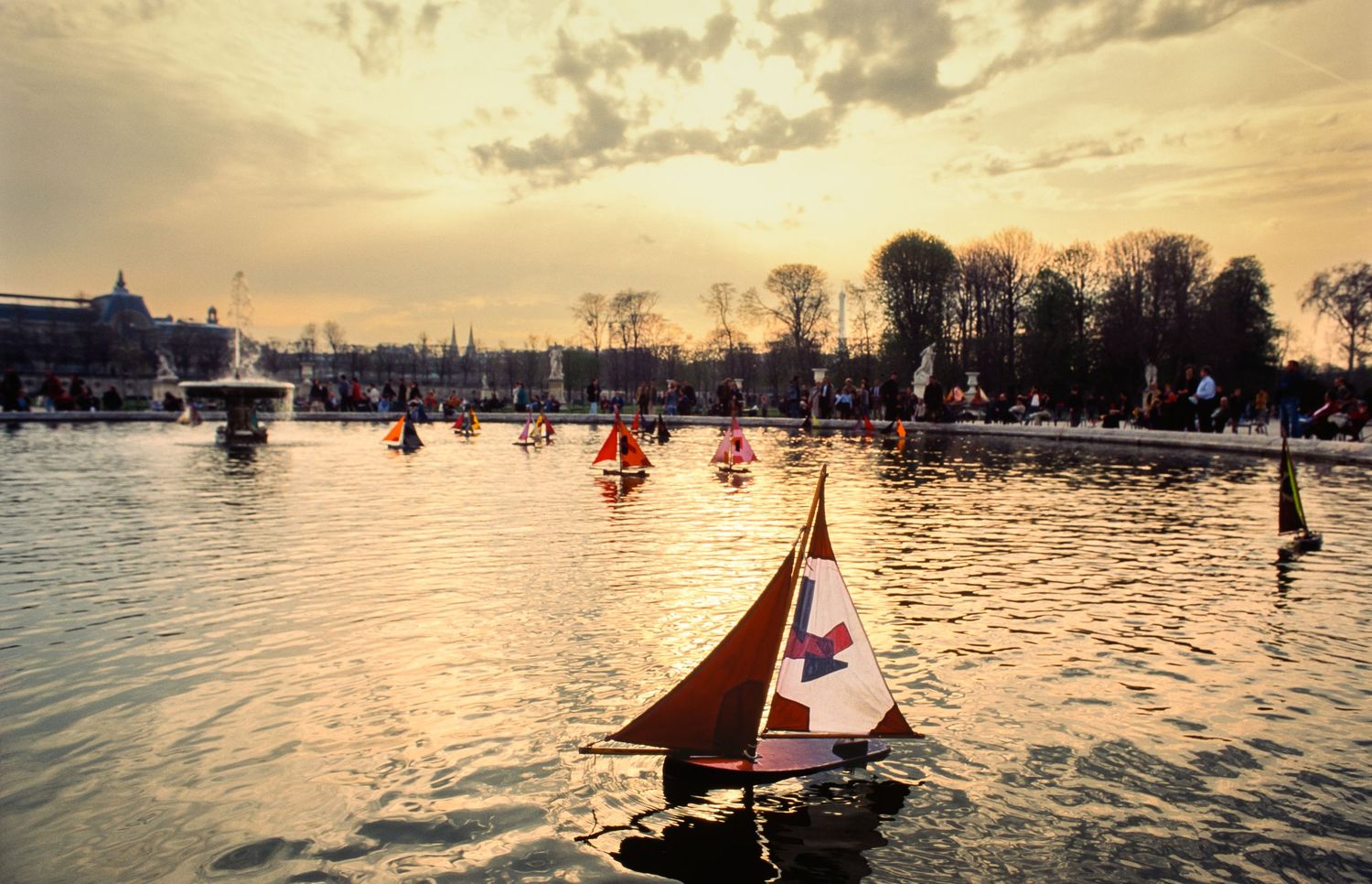 Model sailboats crossing a pond in warm evening light in Paris, France.