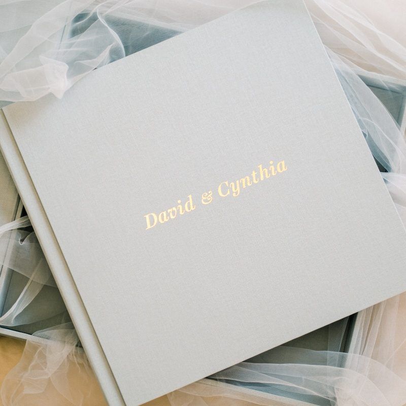 Elegant gray wedding album with gold foil names David & Cynthia, surrounded by soft bridal veil fabric.