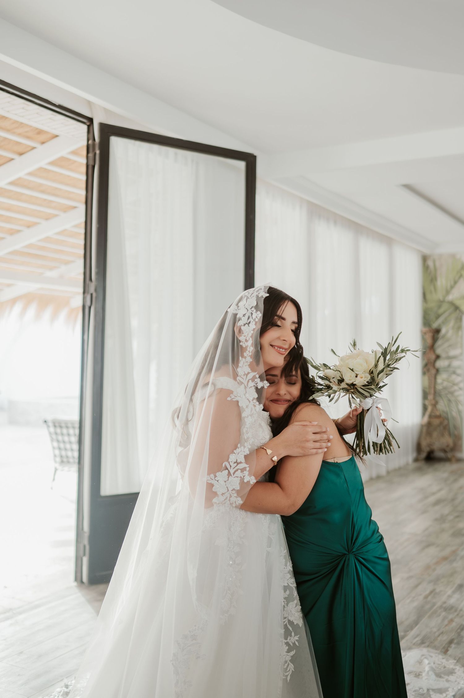 Bride in lace wedding gown embraces woman in green dress while holding white bouquet.