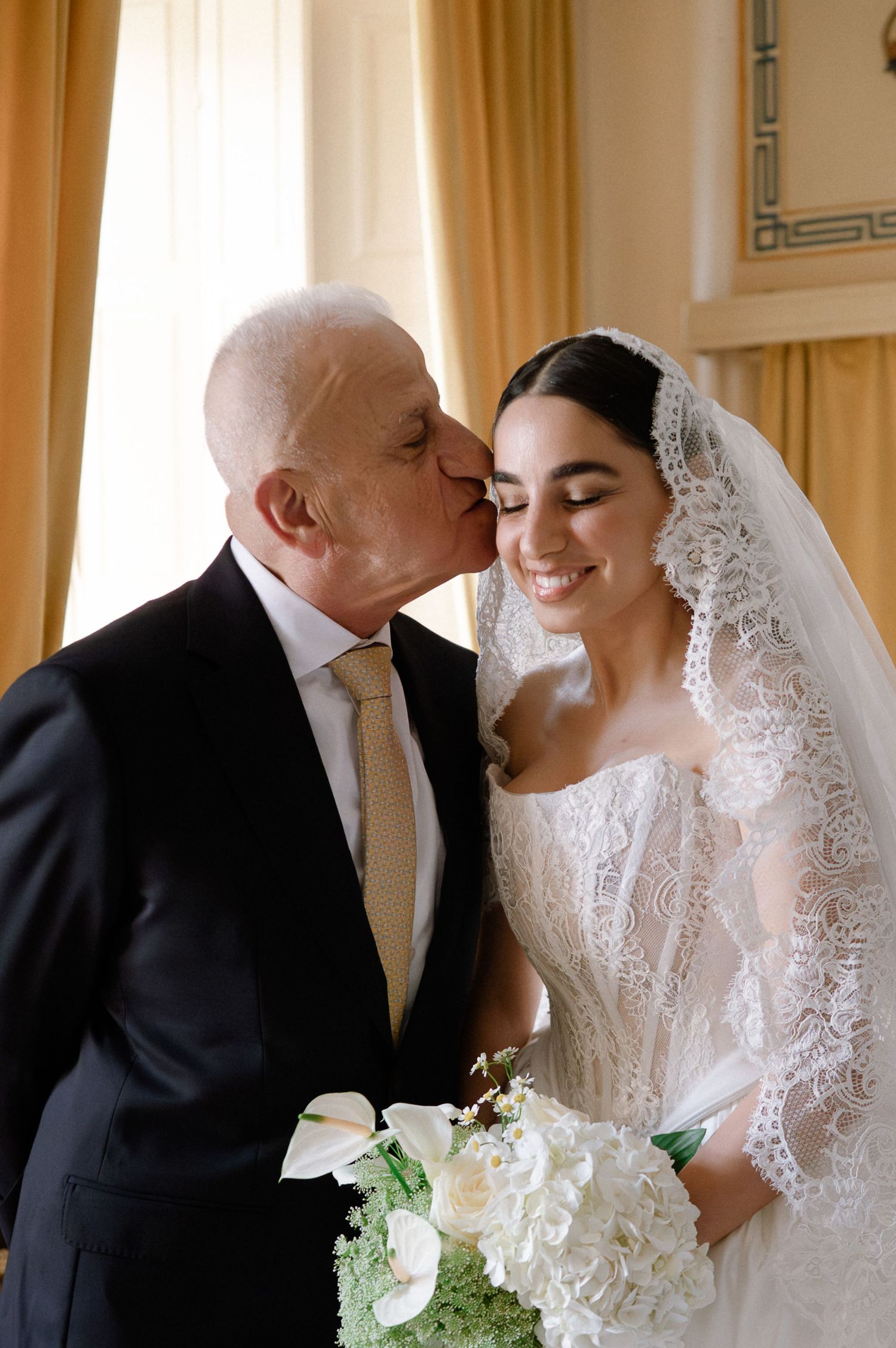 Emotional father kissing bride on forehead as she smiles holding white floral bouquet in elegant wedding dress.