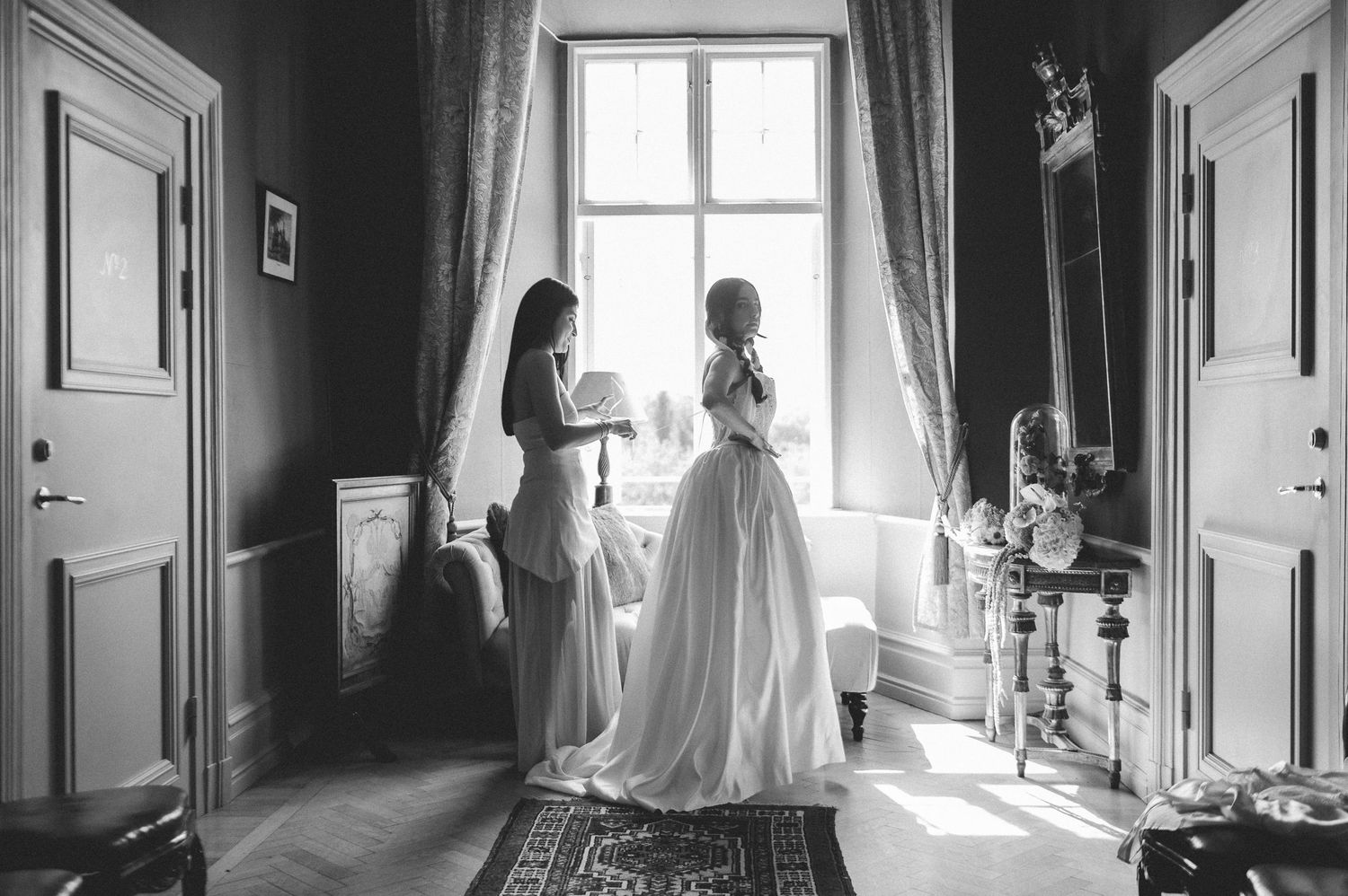 Bride and bridesmaid sharing a moment by window in elegant room, black and white wedding photo.