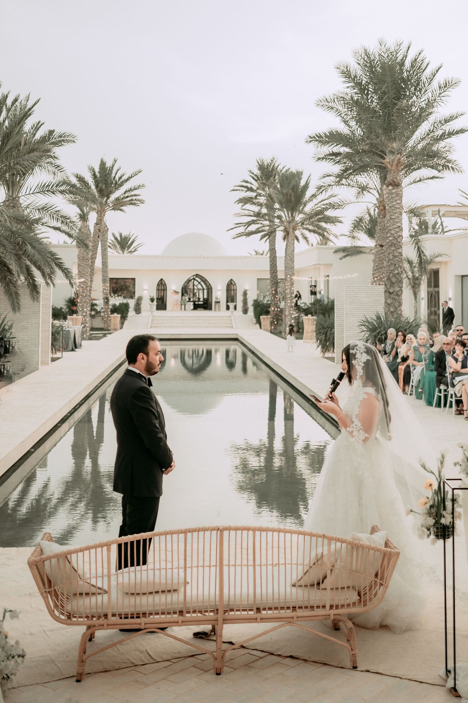 Bride and groom exchange vows at elegant outdoor wedding ceremony beside a long reflecting pool with palm trees.