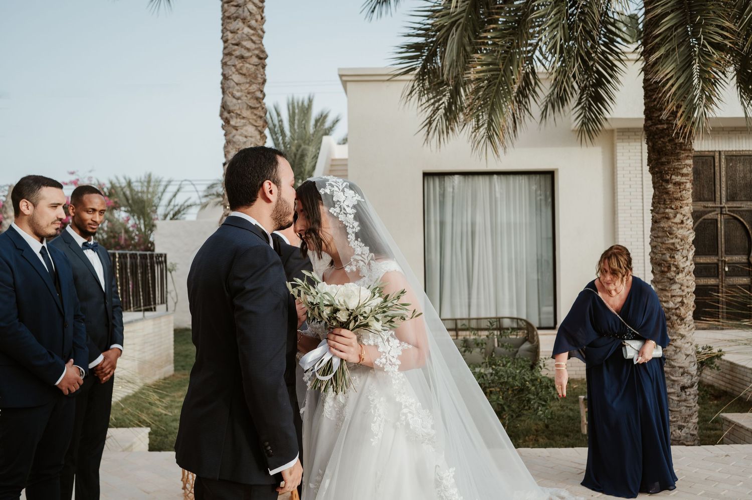 Bride and groom share first kiss at outdoor wedding ceremony with palm trees and guests in formal attire.
