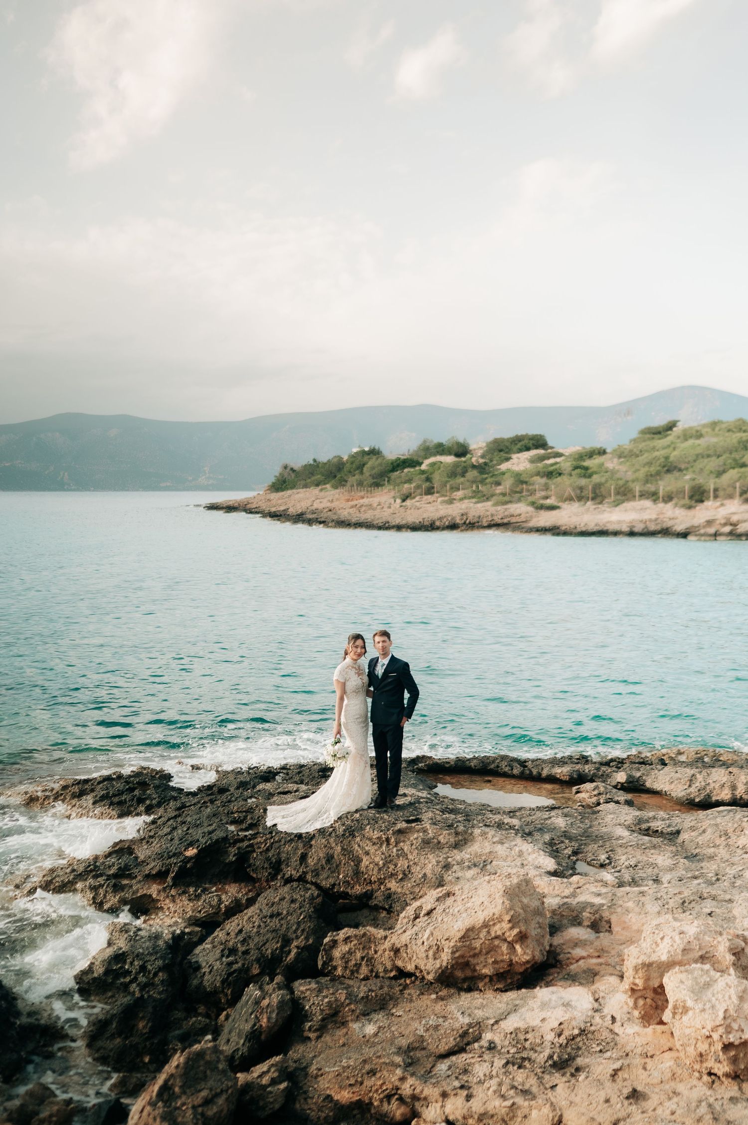 Bride and groom stand on rocky coastal cliffs with turquoise sea and green hills in the background.