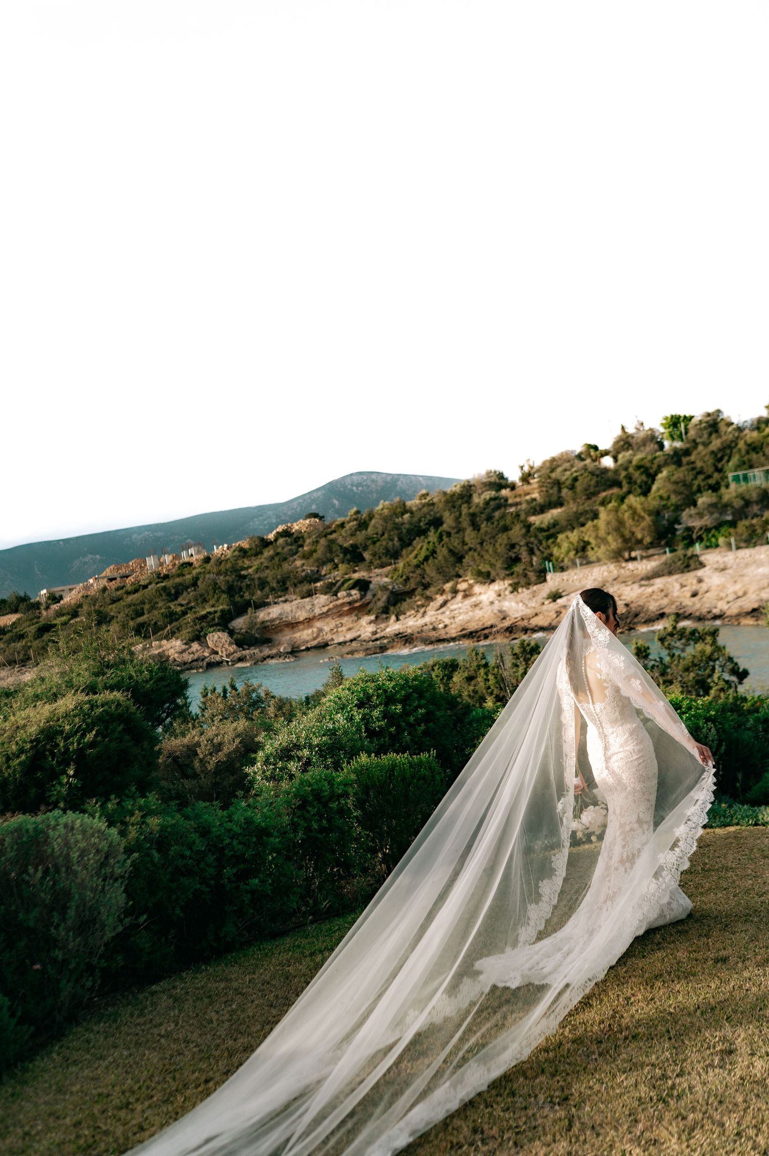 Bride in lace wedding gown with dramatic flowing veil overlooking scenic coastal hillside landscape.