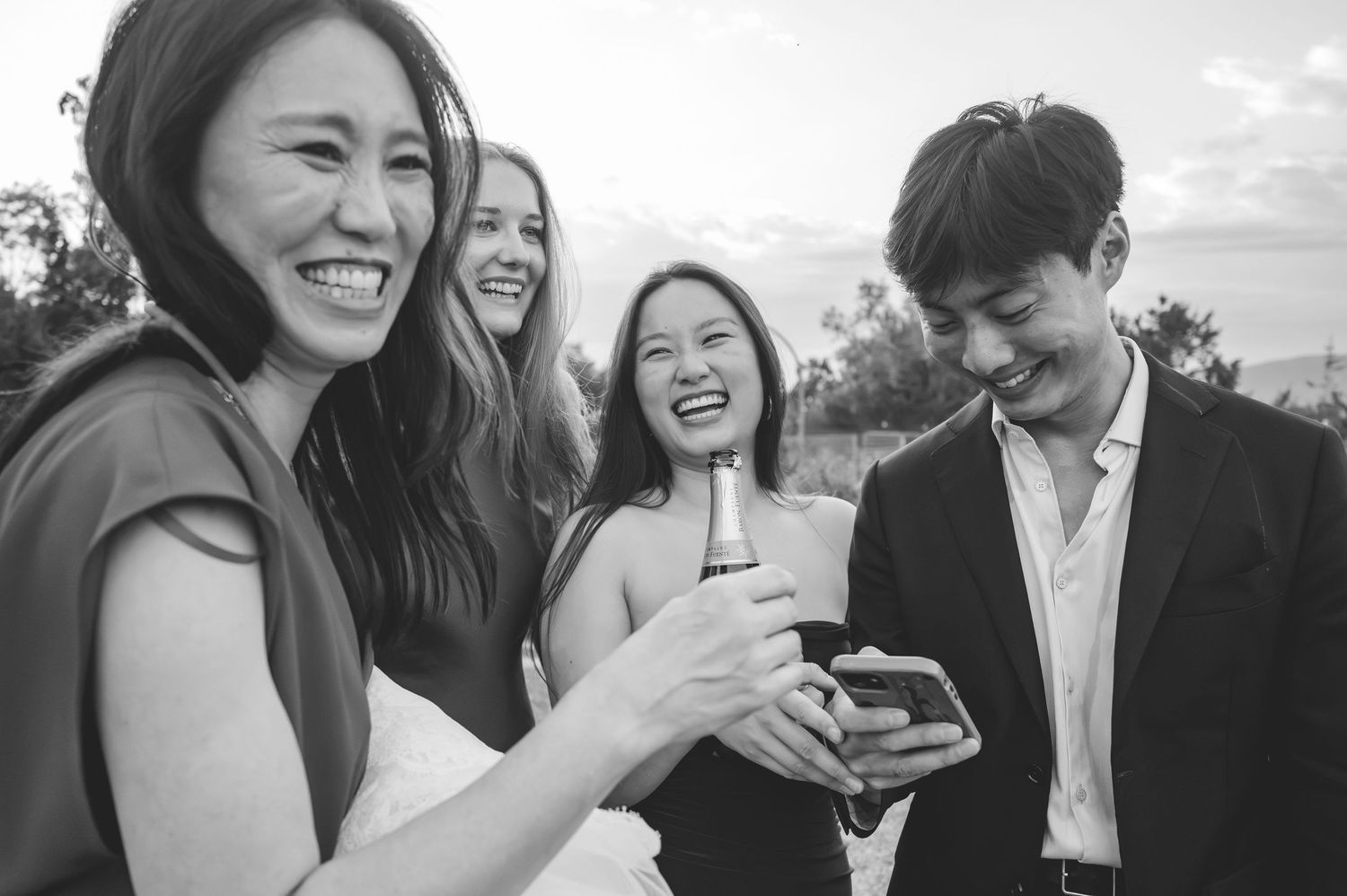 Group of wedding guests laughing and looking at phone during outdoor celebration in black and white photo.