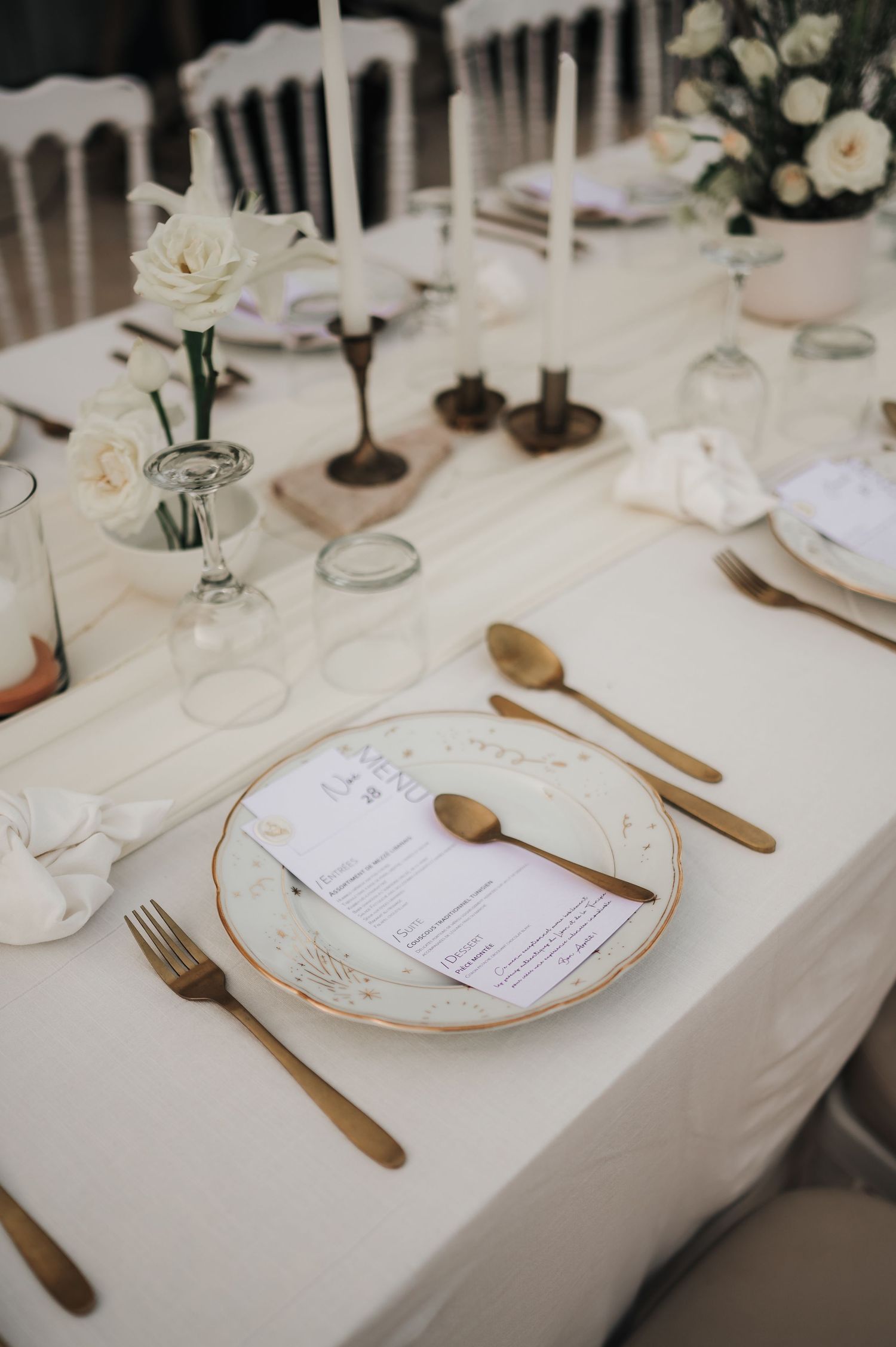 Elegant wedding place setting with gold cutlery, decorative plate, menu card, and white roses on linen table.