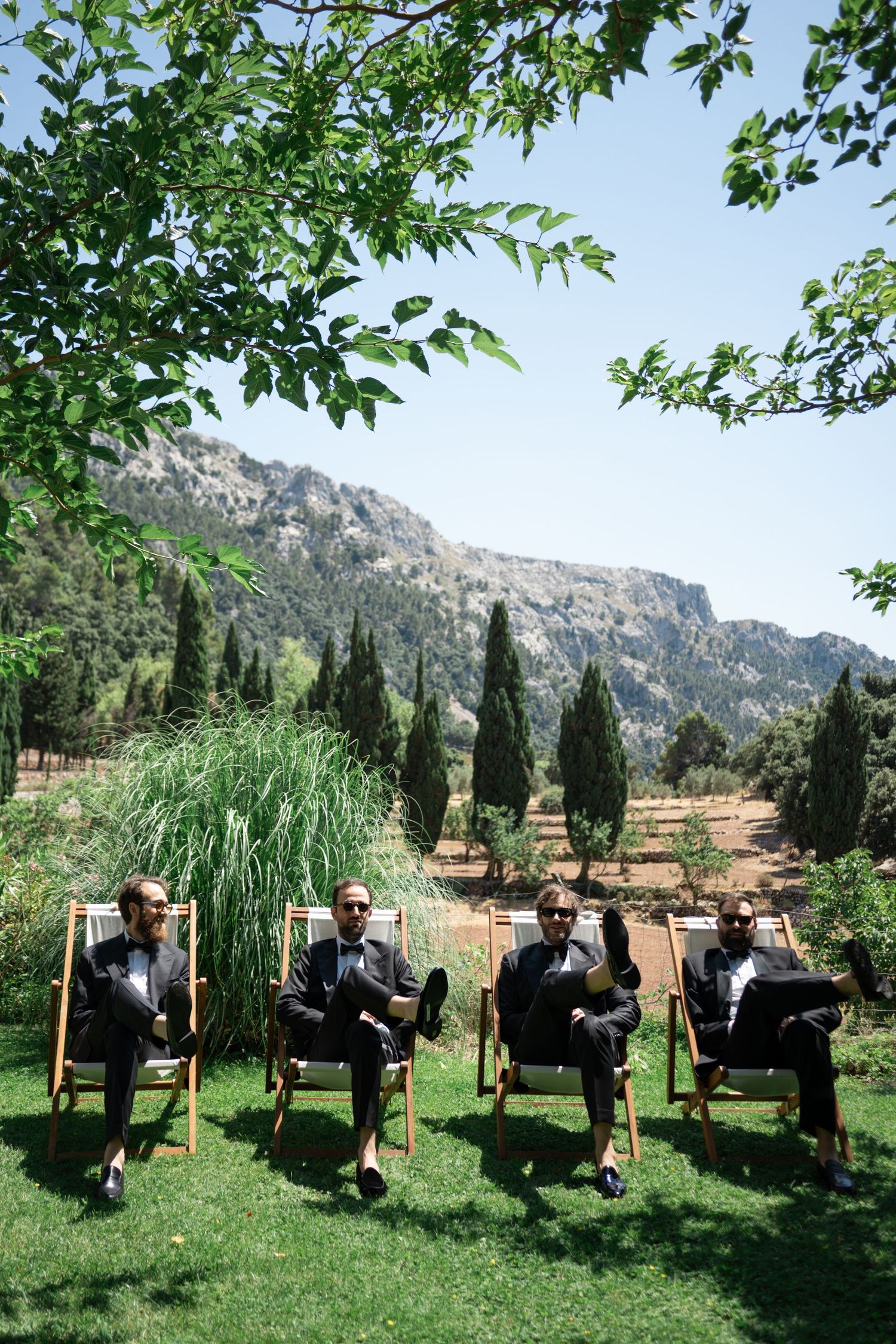 Four groomsmen in black tuxedos relax in deck chairs on a lawn with mountain scenery in the background.