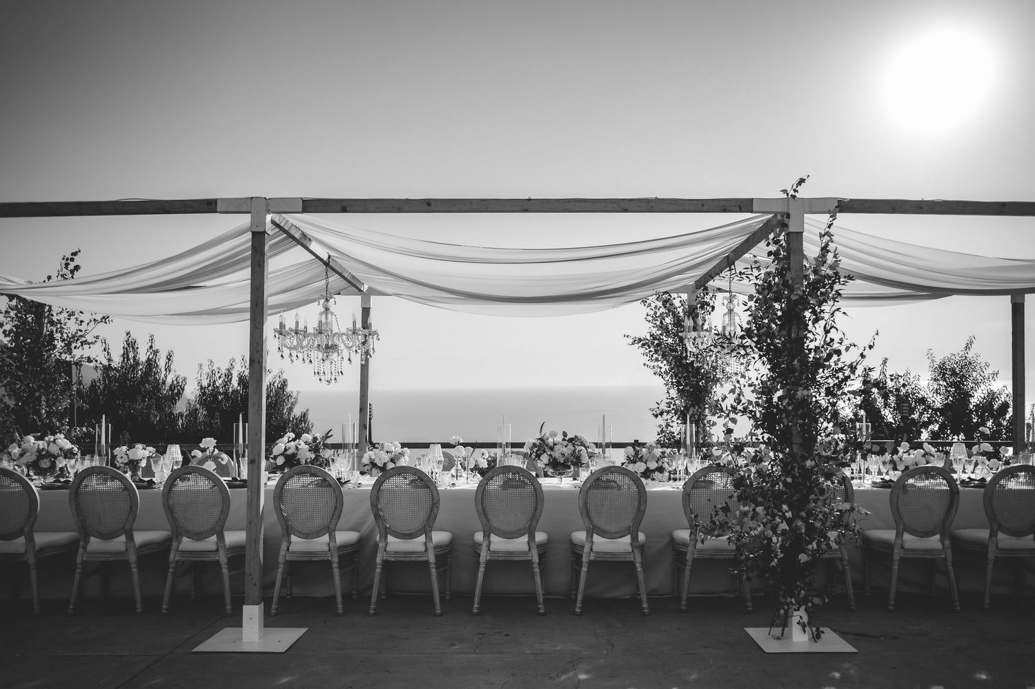 Black and white outdoor wedding reception with long table, draped canopy, floral arrangements, and scenic ocean horizon.