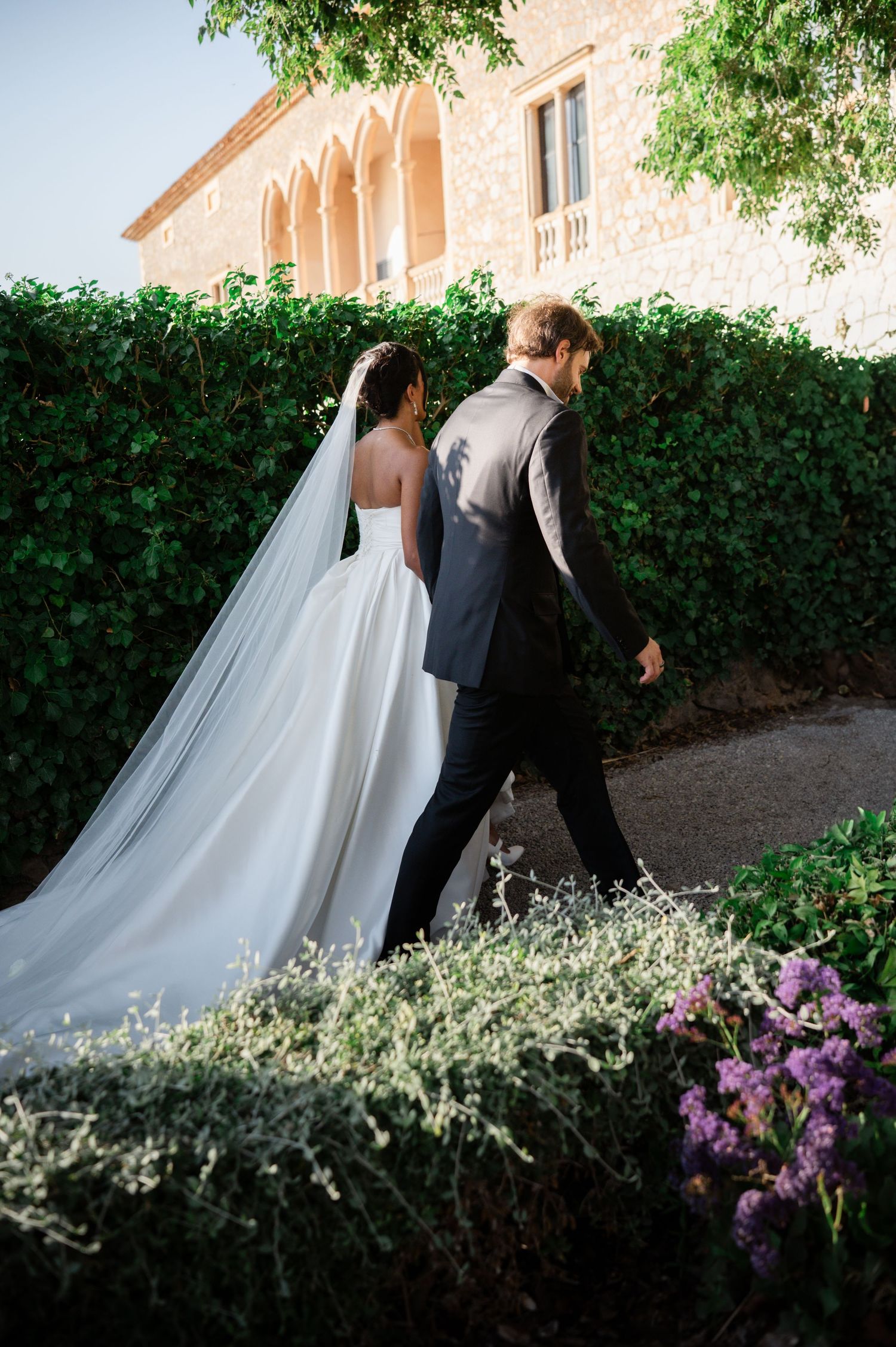 Bride and groom walk past lush ivy-covered stone villa wall with flowing cathedral veil.
