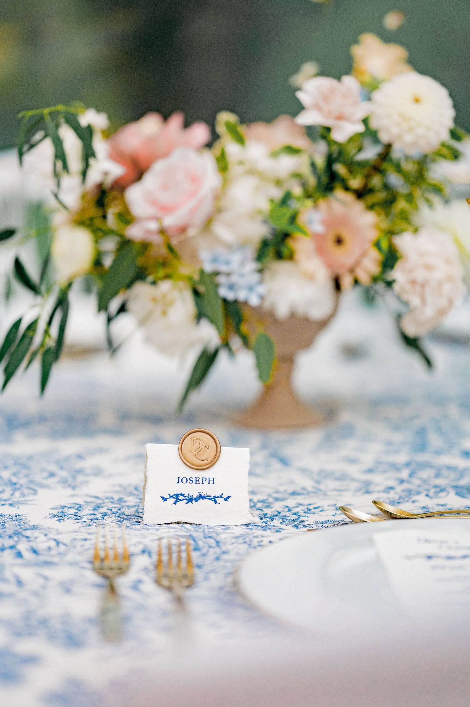 Blue toile tablecloth with gold cutlery, place card reading Joseph and floral centerpiece.