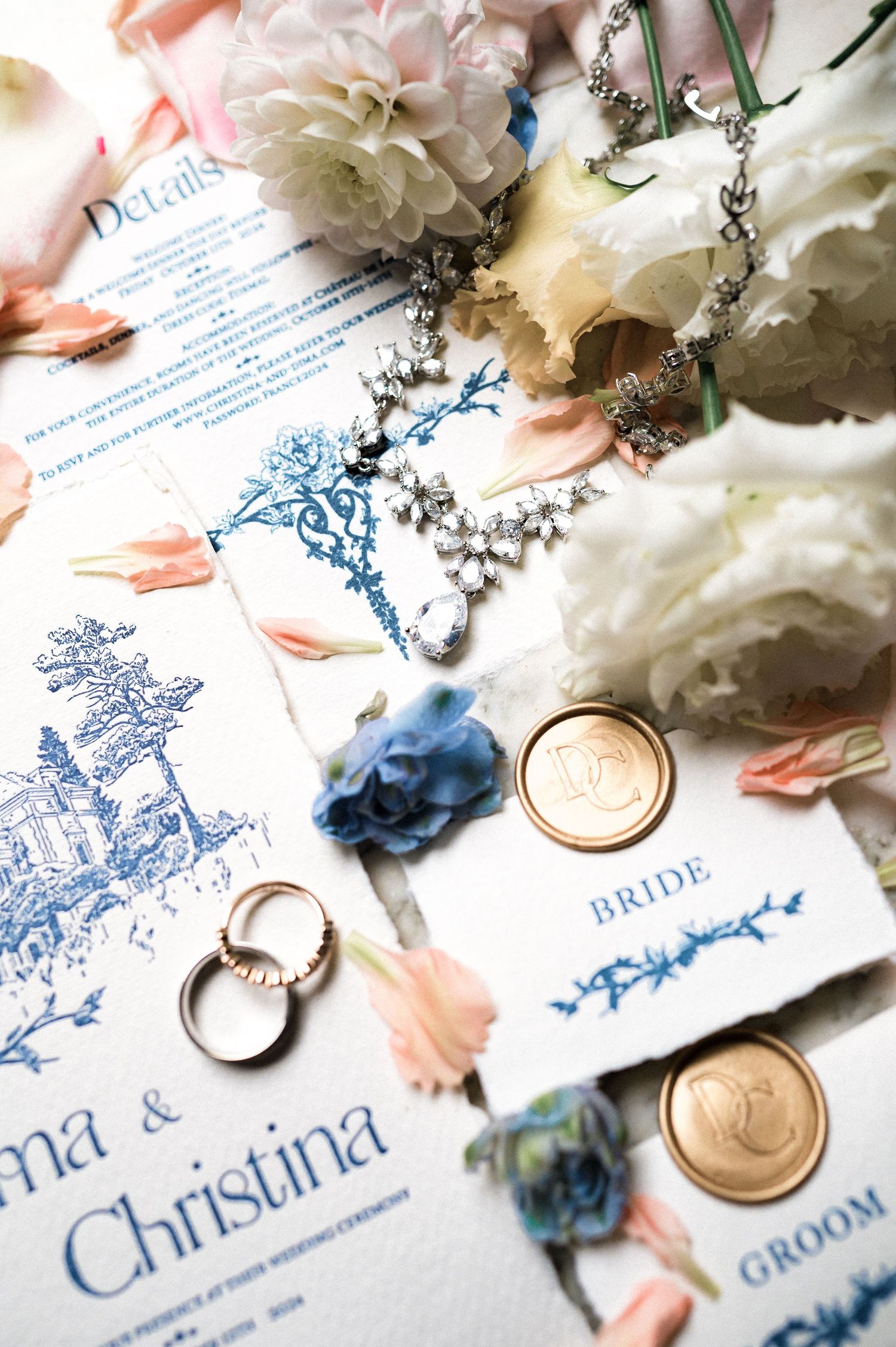 Elegant wedding stationery flat lay with blue toile invitations, gold wax seals, diamond jewelry, and soft floral accents.