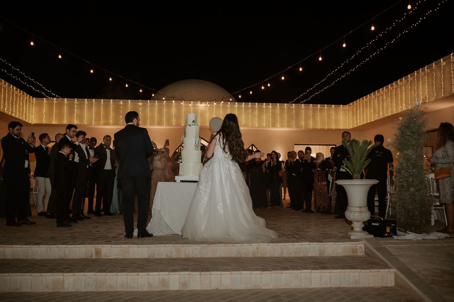 Bride and groom stand before wedding cake at elegant outdoor night reception with string lights.