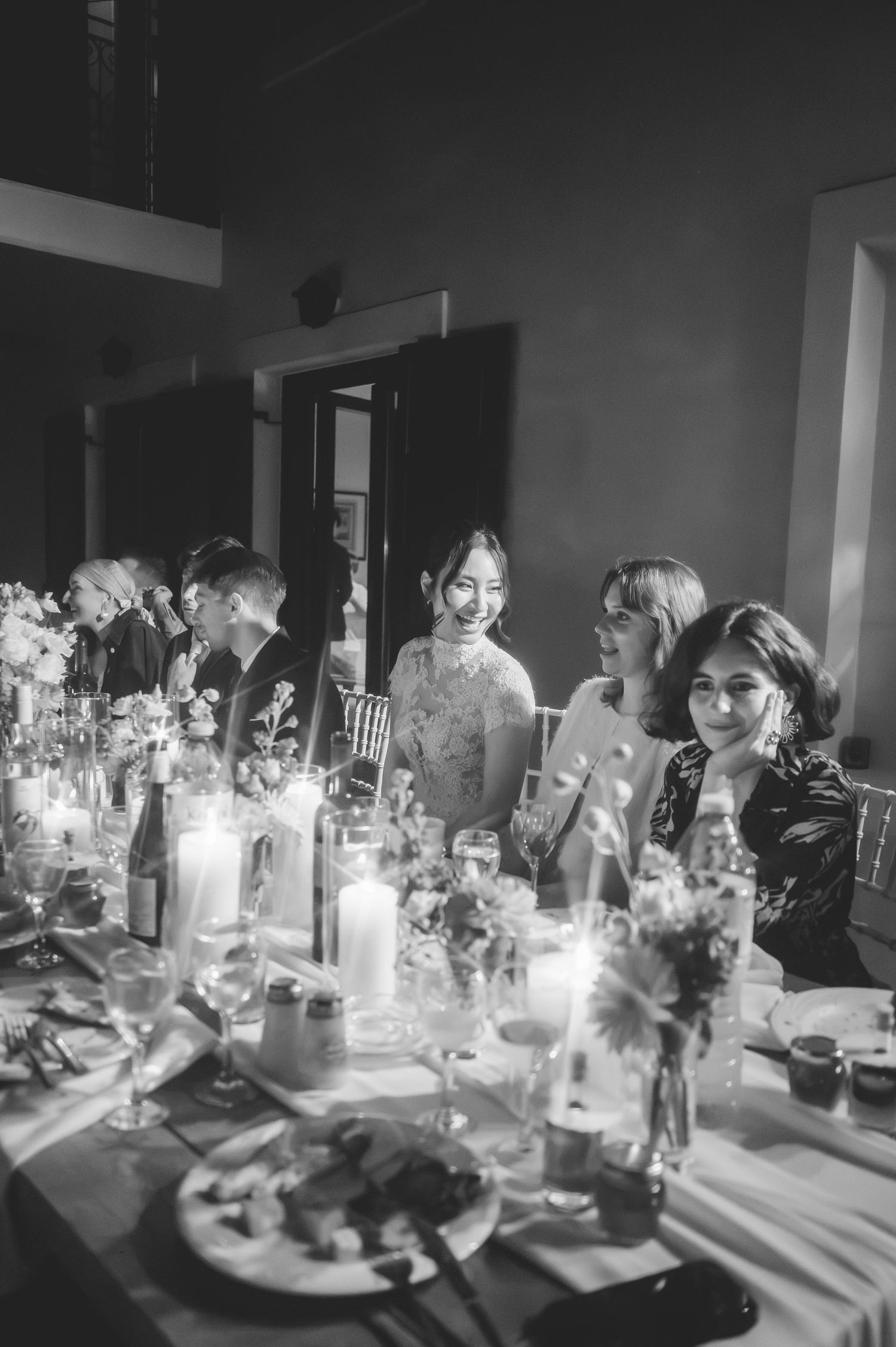 Black and white photo of smiling wedding guests seated at a candlelit indoor reception dinner table.