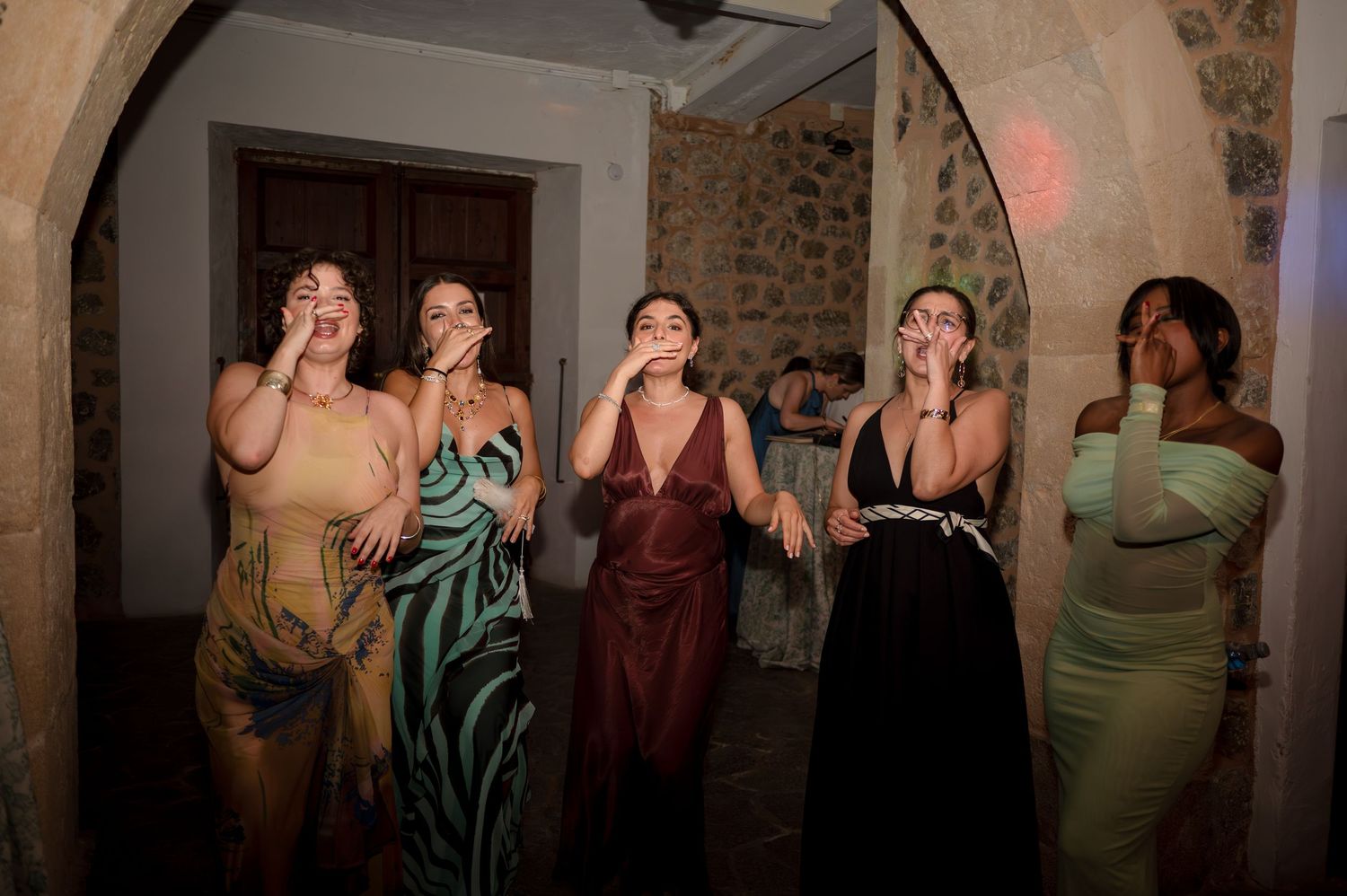 Five elegantly dressed women react with excitement and awe at a wedding reception in a stone archway venue.