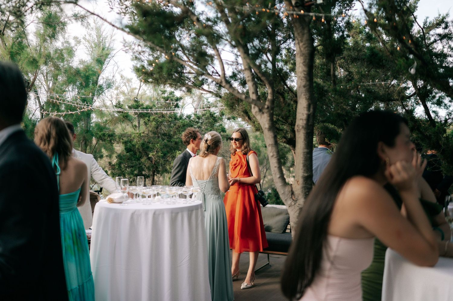 Wedding guests mingling outdoors near cocktail table surrounded by pine trees and string lights.