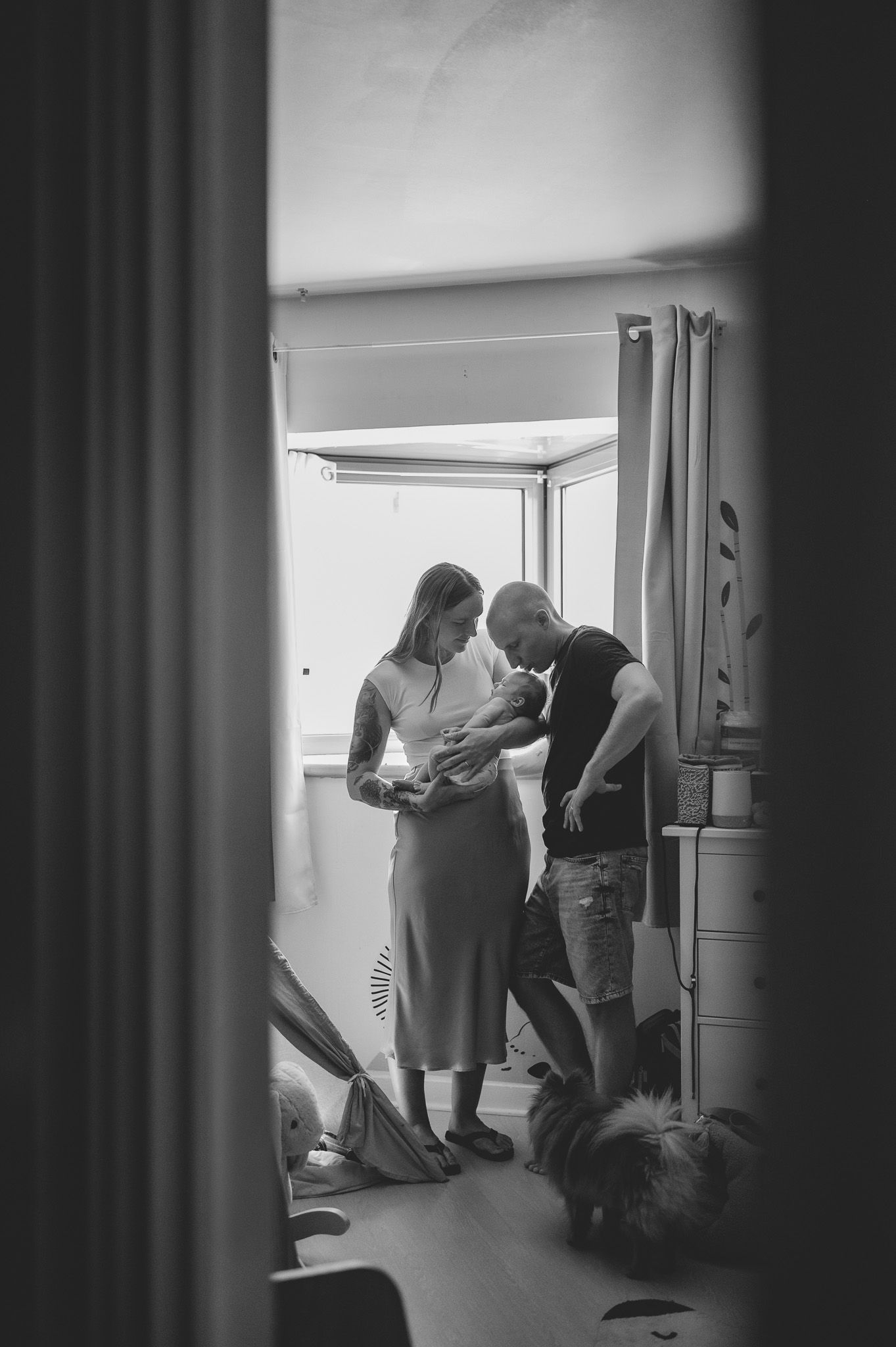 Black and white photo of new parents holding their newborn baby at home with a dog nearby.