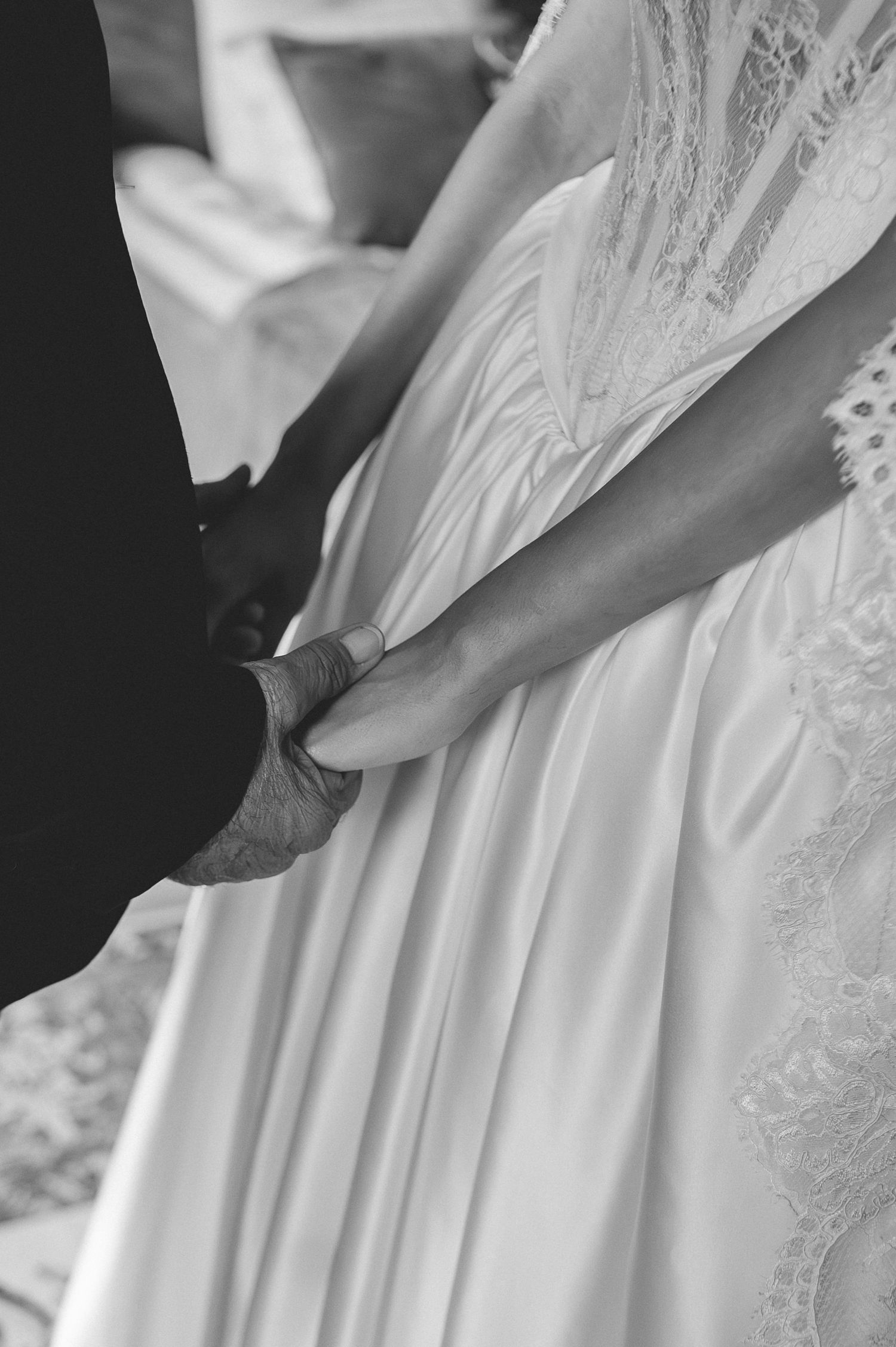 Black and white close-up of father and bride holding hands, bride wearing satin ball gown wedding dress.