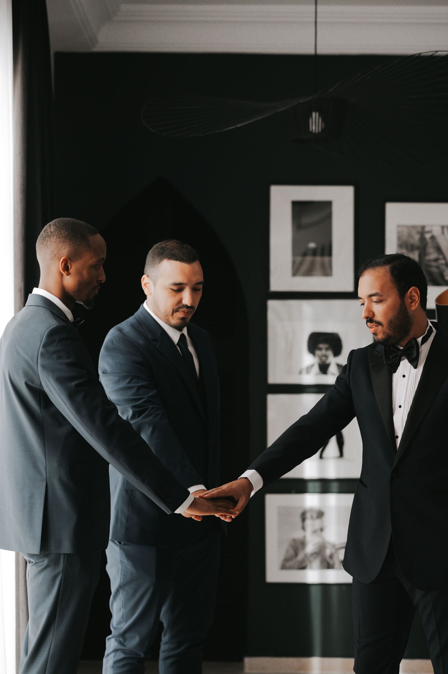 Three groomsmen in dark suits joining hands together in elegant black-walled room.
